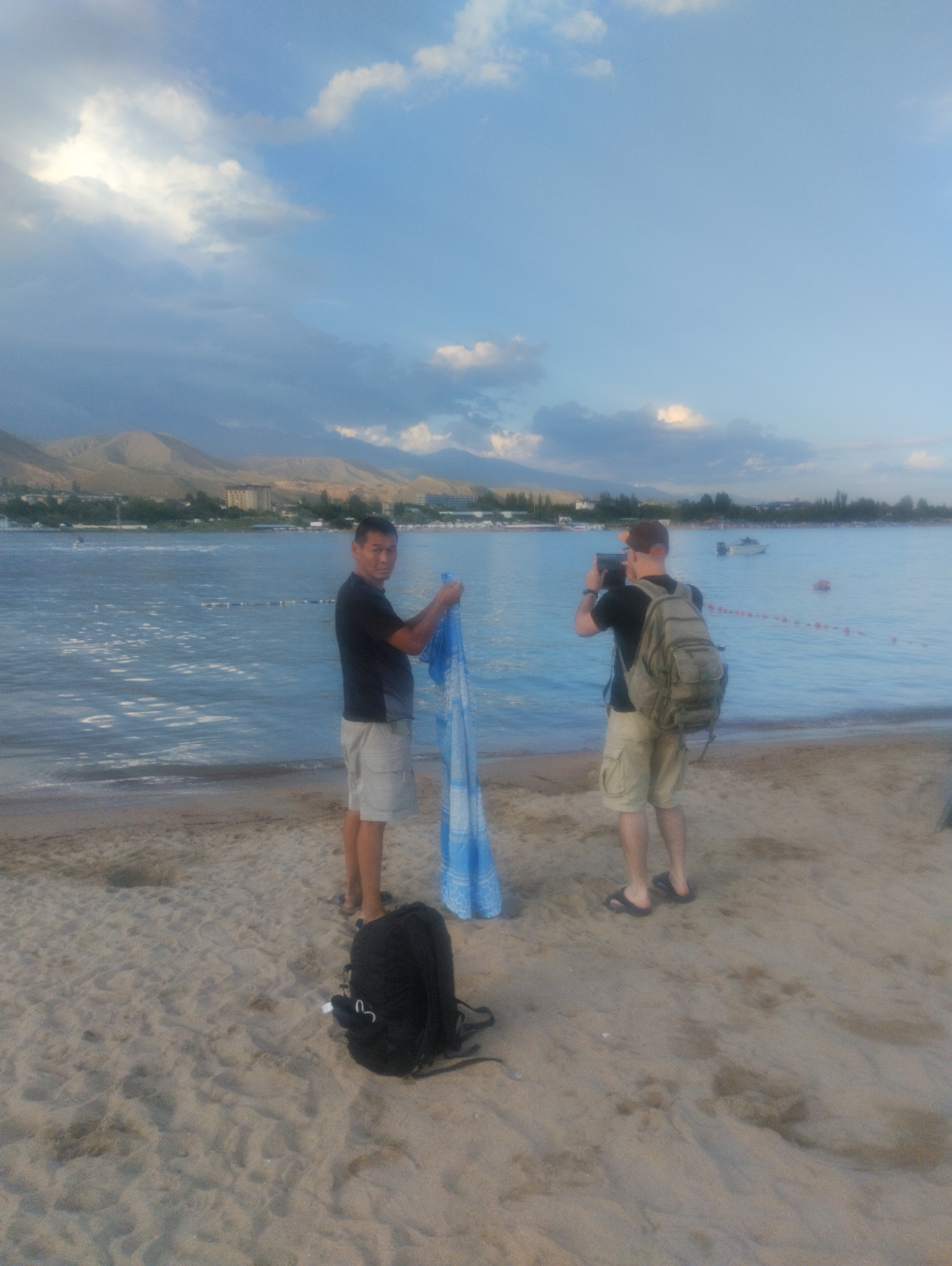 Two men are standing on a sandy beach in Cholpon-Ata, Kyrgyzstan, with a body of water in the background. The man on the left, facing the camera, is wearing a dark t-shirt and light-colored shorts. He is holding a blue and white patterned fabric. The man on the right, with his back to the camera, is wearing a dark t-shirt, light-colored shorts, and a backpack. He is holding a camera or phone and appears to be taking a picture. The water is calm with ripples and some buoys or markers are visible in the distance. Mountains are visible on the horizon behind the water. The sky is cloudy with patches of blue. A black backpack is lying on the sand in the foreground.