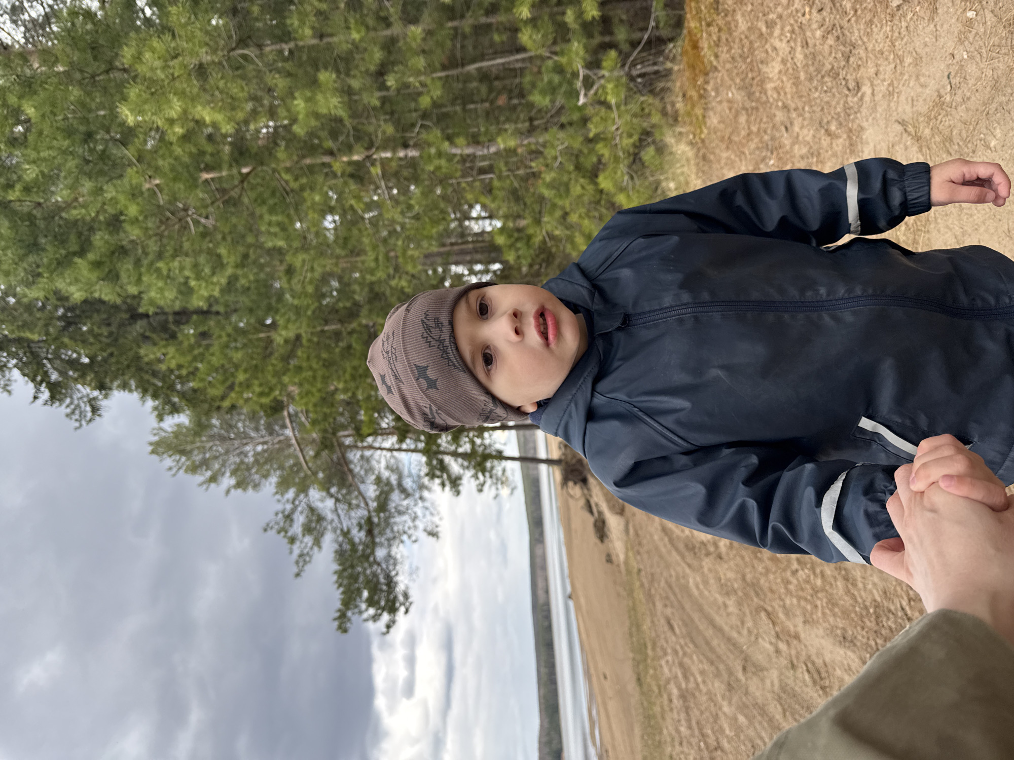 The image features a young child, likely a toddler or preschooler, captured outdoors while looking directly towards the camera with a curious expression. The child is wearing a grey patterned beanie and a dark blue waterproof jacket with a zipper and reflective stripes on the sleeves. Their mouth is slightly open, suggesting surprise or engagement with the person taking the photo.
The child is holding the hand of an adult, of whom only a portion of the hand, lower arm, and a section of an olive/khaki colored garment (possibly pants or a jacket) are visible in the foreground.
The setting is a natural, outdoor environment, specifically a forest edge alongside a body of water. Behind the child and extending to the left of the frame, numerous evergreen trees with green needles create a dense backdrop. To the child's right, a wide expanse of water, possibly a lake or a broad river, is visible, with a sandy or dirt bank sloping down to its edge. The sky above is overcast with grey clouds, indicating cloudy weather during the daytime. The ground in the immediate foreground is unpaved and appears to be sandy or earthy.
The activity appears to be a leisurely walk or pause during an outdoor excursion, with the child seemingly engaged with their surroundings and the adult companion. Given the location context of Chaikovsky, Russia, this scene likely depicts a forested area adjacent to a body of water such as the Kama River or the Votkinsk Reservoir. There is no visible text in the image.