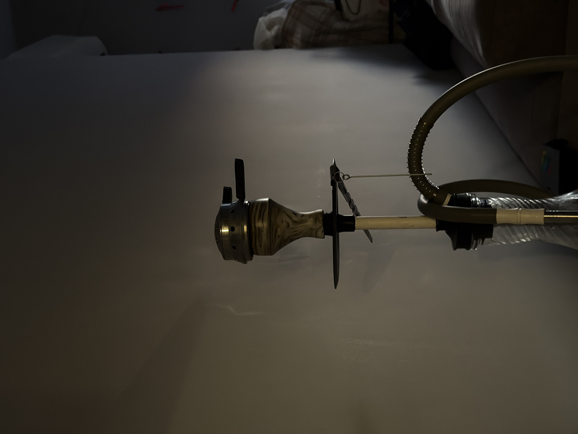 The image displays a close-up, side-view of a hookah hose and stem positioned on a plain, light-colored surface. The hookah stem is made of a light-colored material with a marbled pattern, and it connects to a darker, possibly metallic piece with holes. Extending from the stem is a long, flexible, ribbed hose, colored a muted olive green. This hose curves gracefully upwards and then back down, partially obscuring another section of the hookah apparatus which appears to be a plastic tube. A thin wire or string is attached to a part of the hookah stem and extends towards the hose. The background is dark and out of focus, suggesting an indoor setting, possibly a room with furniture or soft furnishings visible in the upper portion. The lighting is dramatic, with a strong light source casting shadows, indicating it might be nighttime or a dimly lit environment. There are no people visible in the image. The overall impression is one of a still life arrangement of a hookah, with emphasis on its form and texture. There is no visible text in the image. The location context of Sirius, Russia, does not offer specific visual cues within the image itself.