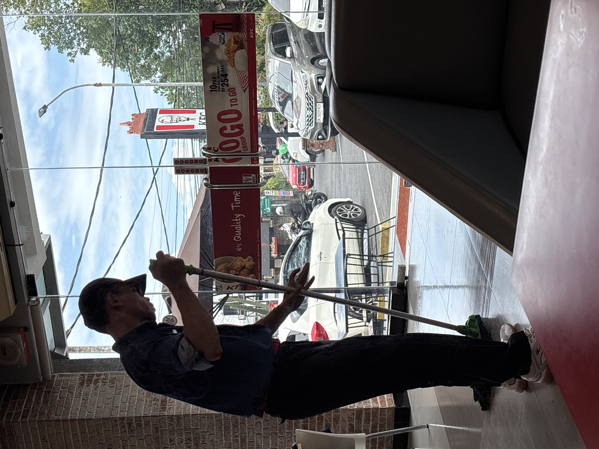 An employee wearing a dark blue shirt, dark pants, and a baseball cap is mopping the tiled floor inside a KFC restaurant in Denpasar, Indonesia. The employee is using a green mop with a long handle. Through the glass storefront, a street scene is visible, including cars, motorcycles, a KFC sign, and a street lamp. The sky is partly cloudy. The interior of the restaurant includes brick walls and tables and chairs, some of which are visible through the glass. A dark gray sofa is in the foreground. The time of day appears to be daytime.