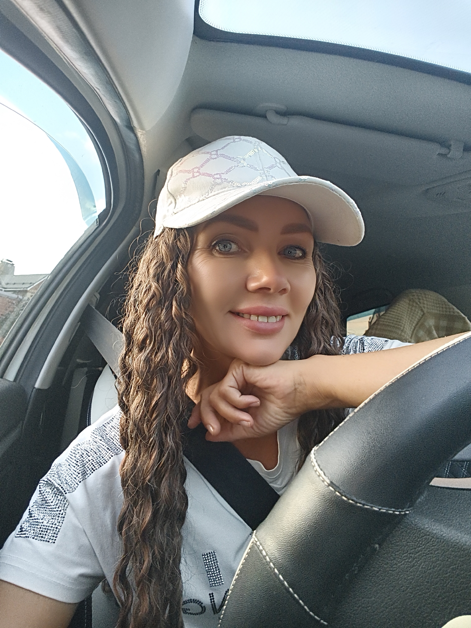 A medium shot captures a woman with long, curly brown hair and blue eyes wearing a white baseball cap and a white t-shirt with sequined details. She is sitting in the driver's seat of a car, her left arm resting on the steering wheel. The woman is smiling and looking at the camera, with her chin resting on her hand. The car interior, including the dashboard, steering wheel, and a seatbelt, is visible. The background shows a glimpse of an urban environment with buildings and a bright sky. The lighting suggests it is daytime.