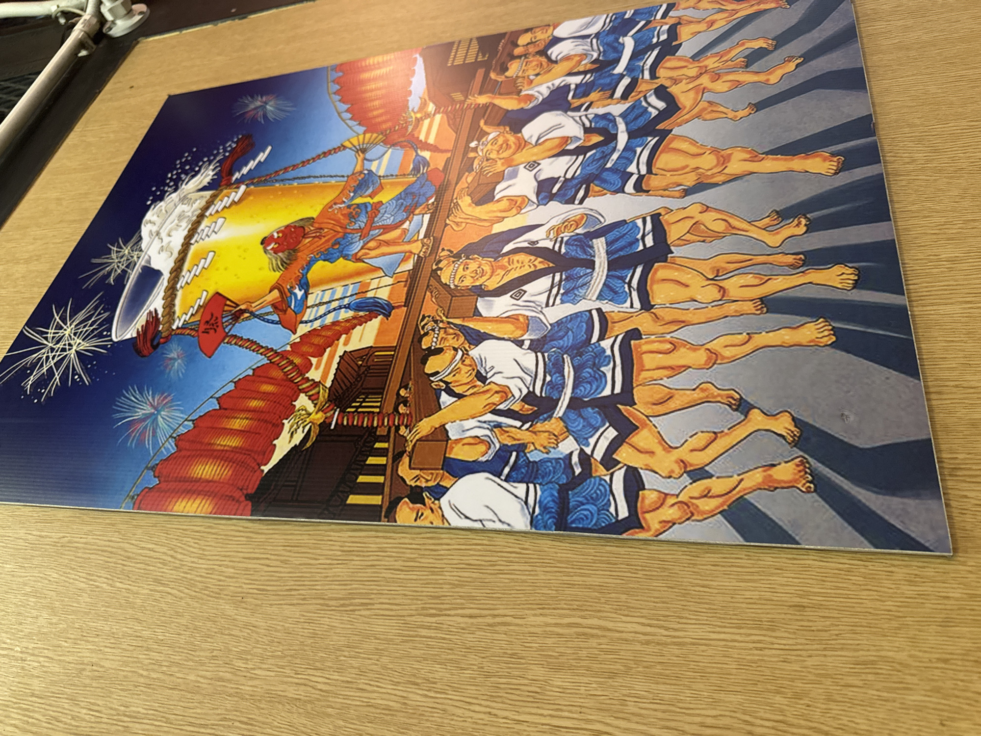 This image displays a vibrant, stylized illustration depicting an energetic night festival scene. Numerous men, rendered in a comic-like art style, are shown participating in what appears to be a parade or a communal event. They wear traditional Japanese festival attire, including white happi coats, blue patterned shorts, and bare legs, exuding determination and joy.
The main activity centers around a large, golden, cylindrical object, resembling an oversized beer mug or sake barrel, from which a frothy white substance overflows. The men are arranged in lines, seemingly pulling ropes attached to a large, ornate float or structure that includes a traditional Japanese building facade adorned with red lanterns. In the upper left, a figure in an orange and blue costume with a red mask holds a large red fan.
The setting is outdoors at night, indicated by the dark blue sky dotted with colorful fireworks. The architecture and traditional clothing strongly suggest a Japanese cultural event. Visible text includes a stylized Japanese character on a red banner, appearing to be "大" (dai, meaning "great"), and some obscured, stylized text on the large cylindrical object. The overall atmosphere is lively and celebratory, depicting a moment of intense, communal festivity.