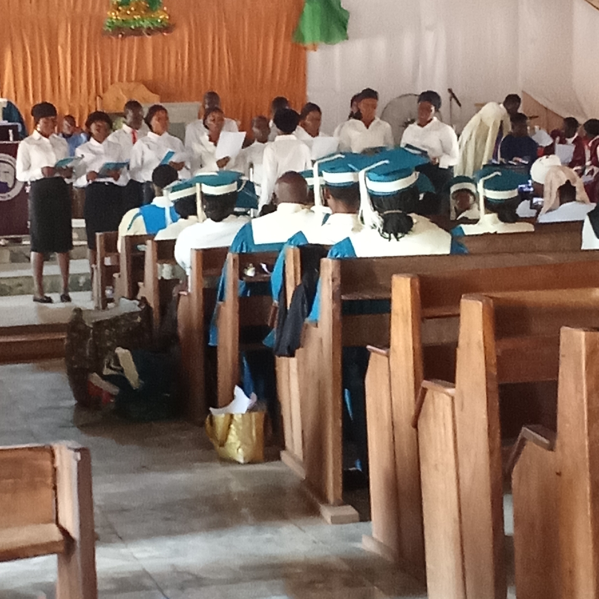 A photograph depicts a church choir in Akwanga, Nigeria. The choir is divided into two sections, one standing and one seated. The standing members, predominantly women, are dressed in white shirts and dark skirts, holding sheet music. The seated choir members wear blue and white gowns with matching caps. They are seated in wooden pews. The background features an orange curtain with a green decoration above it. The church interior appears simple with a concrete floor. Other objects in the scene include a gold-colored bag and other items under a pew. The overall scene suggests a church service or performance.
