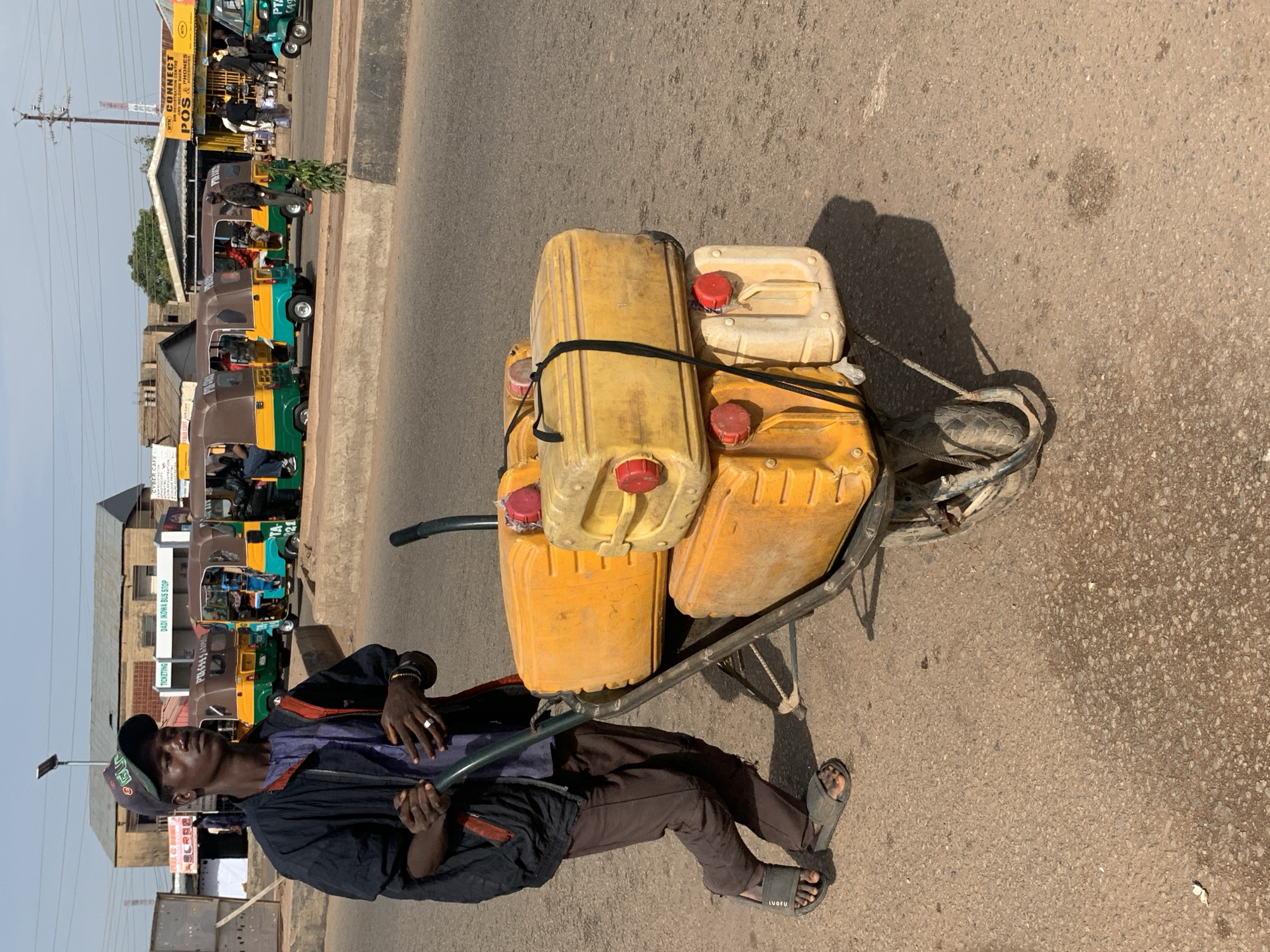 In Gura Top, Nigeria, a man wearing a dark jacket, blue shirt, and brown pants, with a baseball cap on his head, is pushing a wheelbarrow. The wheelbarrow is loaded with four yellow jerrycans, two large and two smaller ones, secured with black straps. The man is walking on a paved road. In the background, there are several colorful auto-rickshaws, buildings, and a few other people. The scene appears to be a busy street or market area.