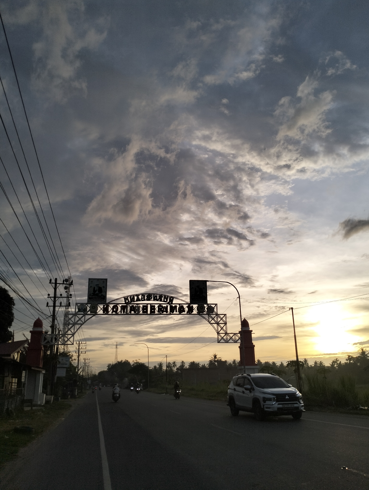 The image shows a road in Tingkeummanyang, Indonesia, under a cloudy sky at sunset. The road leads towards the sun, with a white car driving on the right side. An arched structure spans the road, bearing the inscription "Kawasan Komateerima" in stylized letters. A tower-like structure is on either side of the road where the arch structure is. There are power lines along the left side of the road. Several motorbikes are visible further down the road. Palm trees are visible on the right side of the road in the background.