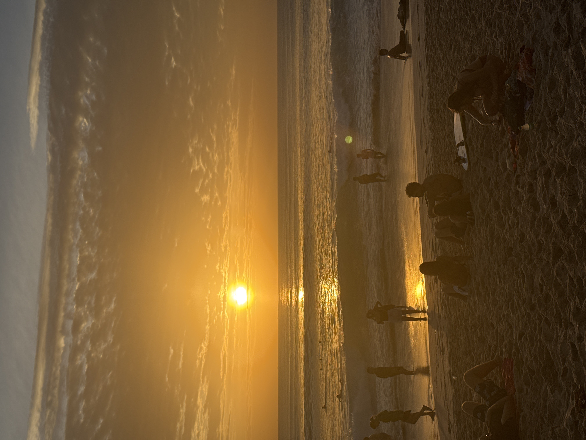 A vertical, low-angle shot captures a beach scene at sunset. The sun, a bright orange orb, is positioned slightly left of the frame's center in the sky, casting a golden glow over the clouds and ocean. The water exhibits gentle waves that break onto the sandy shore. Several people are visible on the beach and in the water. In the foreground, on the sand, are individuals sitting or lying down. Further back, near the water's edge, more people are standing and wading. In the ocean, a few individuals are swimming or surfing. The overall ambiance is one of a serene evening at the coast.