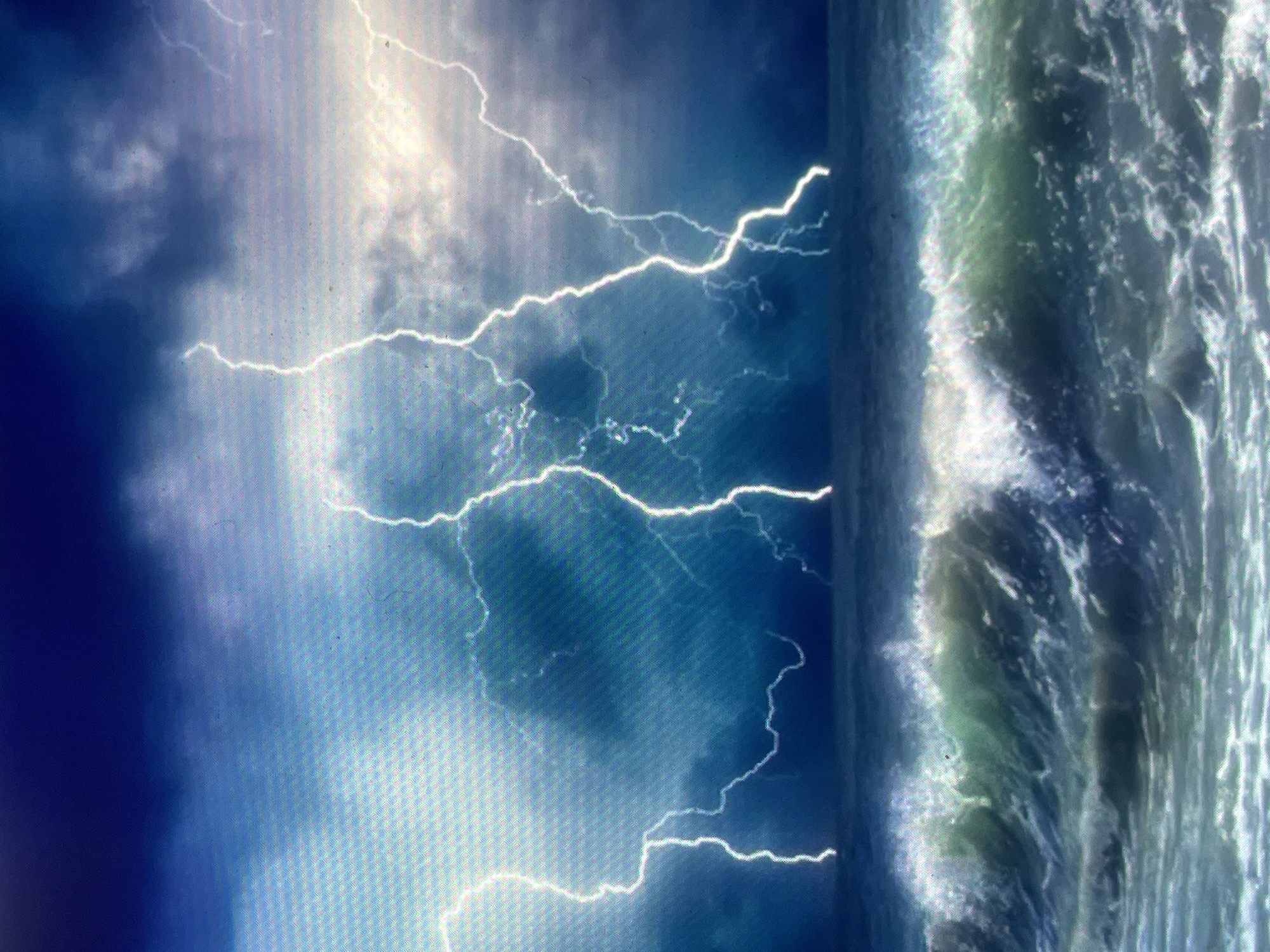 The image, captured from a digital screen and exhibiting a prominent pixel grid, depicts a turbulent open ocean under a severe lightning storm. It does not show the inland city of Zelenyi Hai, Ukraine. The sky is dominated by dark, heavy cumulonimbus clouds, ranging in color from deep indigo to dark gray. Multiple bright, jagged white lightning bolts descend from the clouds, with one particularly intricate and branched strike centrally positioned, reaching towards the ocean surface. The sea below features large, breaking waves with foamy white crests against deep blue-green water, indicating rough conditions. No people, vessels, or artificial structures are present. The scene conveys a powerful and dramatic natural weather event.
