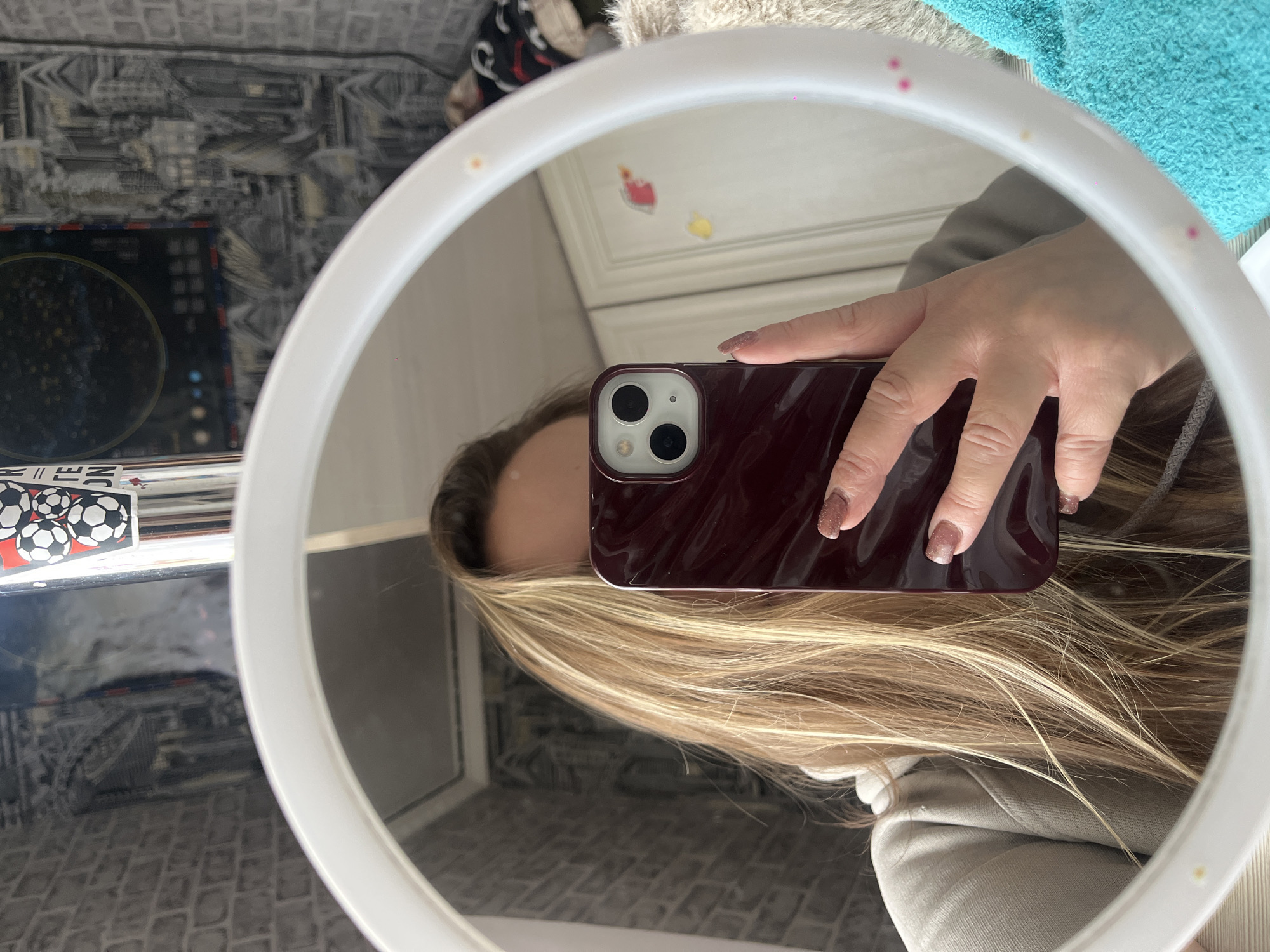 The image captures a person taking a mirror selfie in an indoor setting. The central focus is a circular, white-framed mirror, likely a ring light, reflecting the scene.
A person's right hand, with manicured, sparkling brownish-nude nails, holds a dark red or burgundy-cased smartphone (appearing to be an iPhone with two camera lenses) directly in front of their face. Long, light blonde hair cascades around the phone and hand, and a partial view of the person's forehead is visible above the phone. The person appears to be wearing a light-colored sweatshirt or jacket.
The white frame of the mirror features small, scattered pink glitter-like dots.
The background reflected in the mirror offers glimpses of the room. Behind the person's reflected head, two small, decorative stickers—one red, heart-shaped, and one yellow, abstract—are visible on a light-colored wall or furniture. To the left of the mirror's reflection, there's a dark object that looks like an electronic device with glowing blue and white lights. Below this, a stack of items shows a black and white soccer ball pattern, and some illegible white text, possibly "N TE," is faintly visible. A textured grey and white patterned fabric, possibly a bedspread or blanket, occupies the lower and left portions of the reflected background. In the upper right corner of the reflection, a fluffy turquoise towel or blanket adds a contrasting color element.
The image is rotated counter-clockwise, giving it an unconventional orientation that highlights the act of a mirror selfie. The lighting is consistent with an indoor environment.