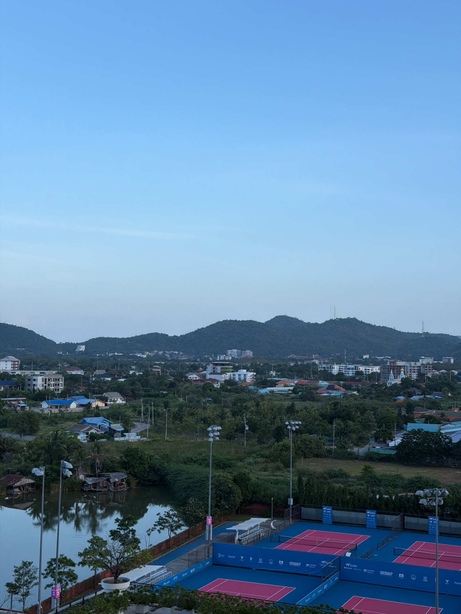 An elevated, wide-angle view captures a serene landscape in Hua Hin City Municipality, Thailand, at either dawn or dusk, with a clear, gradient blue sky dominating the upper portion of the frame. The middle ground reveals a dense panorama of verdant trees interspersed with residential buildings and a distant mountain range. The foreground features a body of calm water reflecting the overcast sky and a cluster of rustic, stilted structures, possibly homes or fishing platforms. Adjacent to the water are several brightly colored tennis courts with blue surfaces and pink lines, equipped with stadium lighting and fencing. The scene is devoid of any visible people or discernible activity.