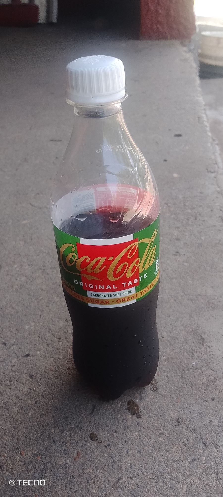 A Coca-Cola bottle with a white cap, containing a dark colored liquid, rests on a concrete surface in Zango-ndaji, Nigeria. The bottle is partially covered in condensation. The label on the bottle features the iconic Coca-Cola logo in red and gold, with green accents. Text on the label indicates "ORIGINAL TASTE" and "CARBONATED SOFT DRINK". Below this, smaller text reads "LESS SUGAR - GREAT TASTE". The concrete surface appears to be outdoors, with some debris scattered on it. A faint logo for "TECNO" is visible in the lower left corner of the image.