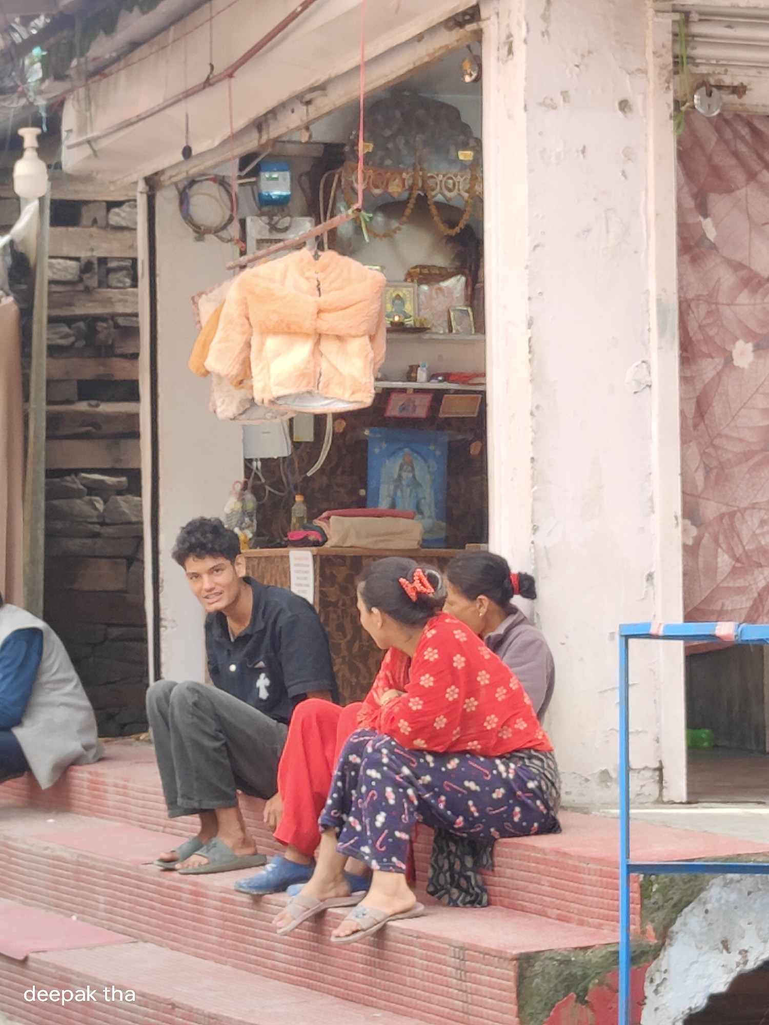 A low-angle, eye-level outdoor shot captures a small shop entrance in Vashisht, India. The entrance has a white facade and is adorned with an orange jacket hanging on display. Behind the jacket, a blue electrical meter is visible. A young man with curly black hair, wearing a dark blue shirt and grey pants, sits on the steps facing the viewer and smiling. Two women are also seated on the steps. The woman in the foreground wears a red top with a floral pattern and patterned pants, while the woman behind her wears a grey top. The steps are made of red brick and show signs of wear, with green moss growing in some areas. The shop appears to be a street-side establishment, possibly a small vendor or stall.