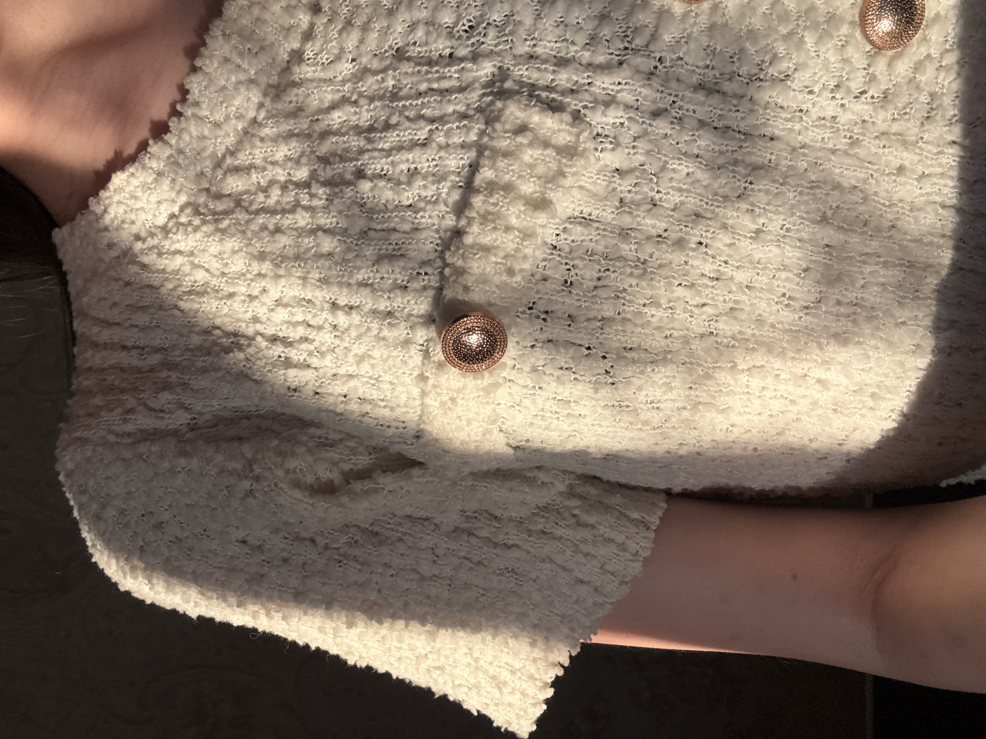 The image shows a close-up view of a person's torso wearing a textured, cream-colored top with short sleeves. The top has visible buttons, likely decorative, with a rose gold metallic finish. One button is prominently featured on the chest, and another is partially visible lower down on the garment. The fabric of the top appears to be a looped or bouclé knit, creating a soft, textured surface. Sunlight is casting strong shadows across the garment, highlighting its texture and shape. A portion of the person's arm is visible on the left side of the frame, showing fair skin. The background is dark and out of focus, suggesting an indoor setting or a shaded outdoor area. There is no identifiable information in the image to associate it with Rayevskiy, Russia.