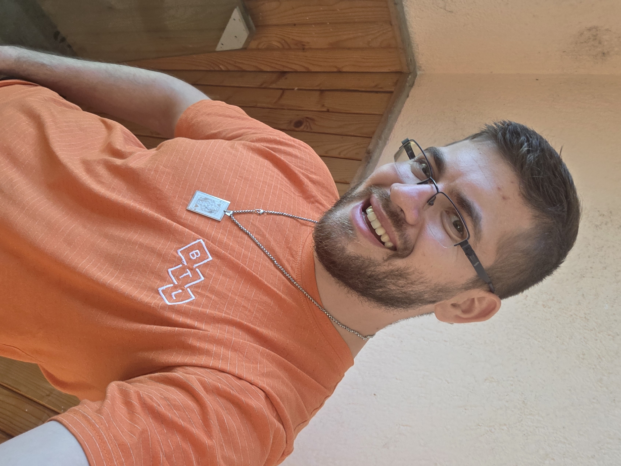 The image is a close-up selfie of a man with brown hair and a beard. He is wearing glasses and an orange shirt with a logo on the front. He is also wearing a silver chain necklace with a rectangular pendant. The background is a wooden wall and a light-colored wall. It is not possible to determine if the photo was taken in Yagodovo, Bulgaria, or to establish any details about the surrounding environment beyond what is visible in the image.