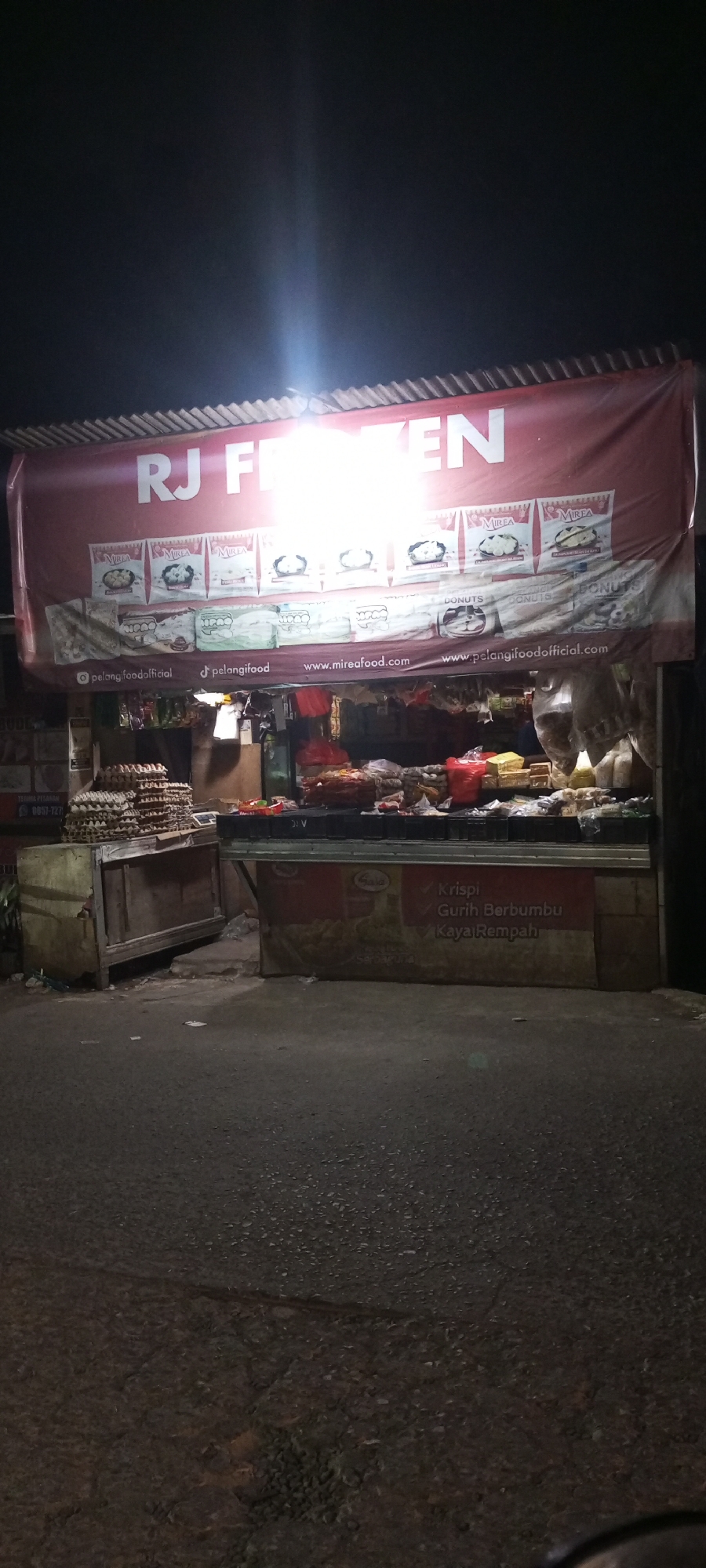 The image shows a roadside food stall at night in Setu, Indonesia. The stall is covered by a red banner that reads "RJ FRozen" in large white letters. Below the banner, there are various food items displayed on shelves. To the left of the stall, stacks of egg cartons are visible. The ground in front of the stall appears to be asphalt, with some debris. The lighting suggests that artificial lights are illuminating the scene, likely from the stall itself and surrounding areas. The overall impression is of a typical street-side food vendor operating after dark.