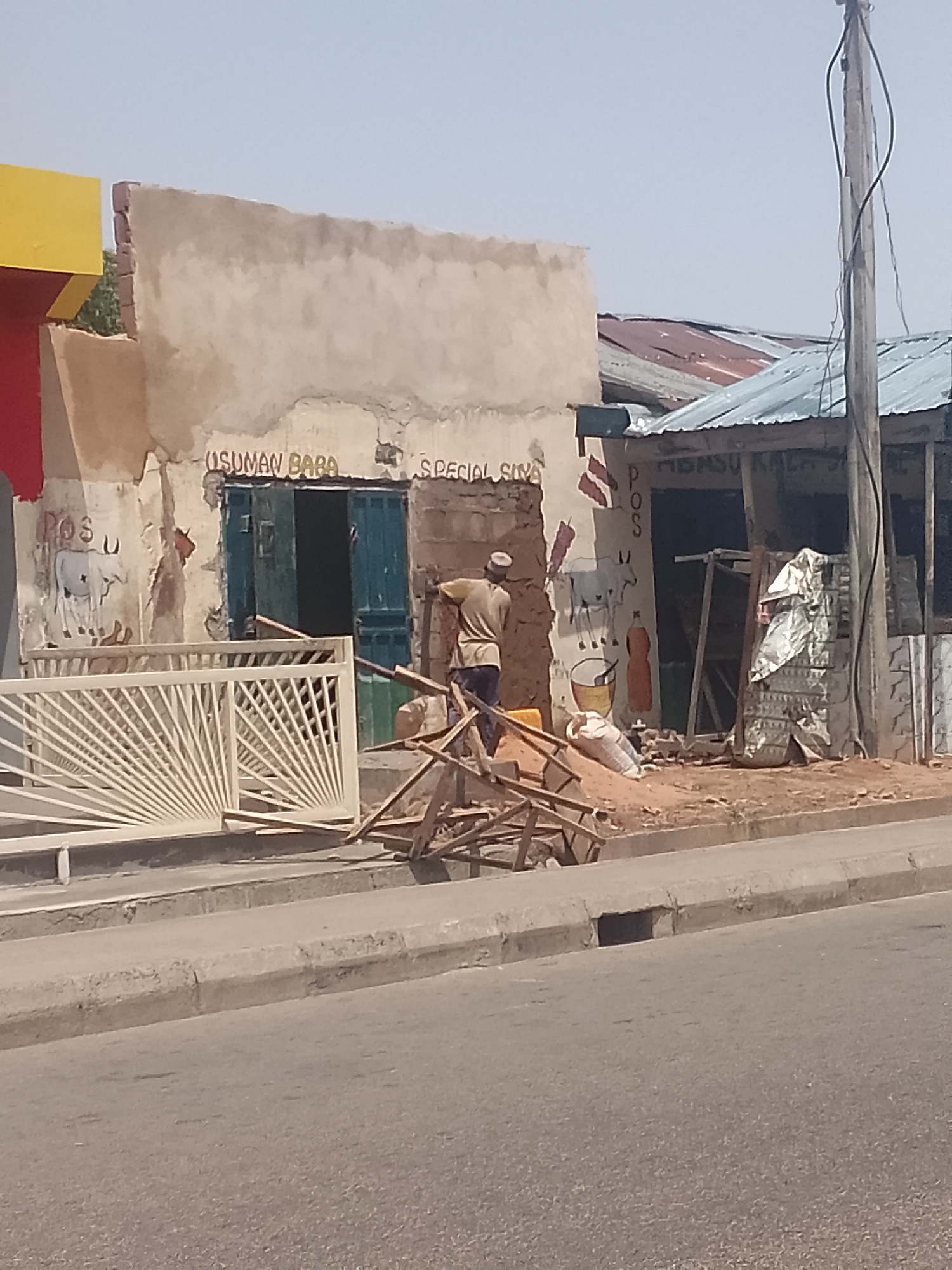 The image shows a street scene with several buildings and a man working on the construction or renovation of a structure.
Buildings: Two buildings are prominent. One has the words "USUMAN BABA" and "SPECIAL SILVA" painted on its facade. The other has "MBASU KALA" written on it. One building has painted drawings on it as well. The buildings appear to be small shops or businesses.
Man: A man wearing a beige shirt and dark pants is working near one of the buildings. He is near a pile of wooden pieces.
Objects: A white metal gate leans against one building. A pile of wooden sticks is on the sidewalk, and sacks and what appears to be construction materials are near the buildings.
Street: A concrete curb separates the sidewalk from the paved street.
Environment: The scene appears to be in an urban or semi-urban area, possibly a small town or city in Unknown, Unknown. The overall impression is one of a developing or low-income area.