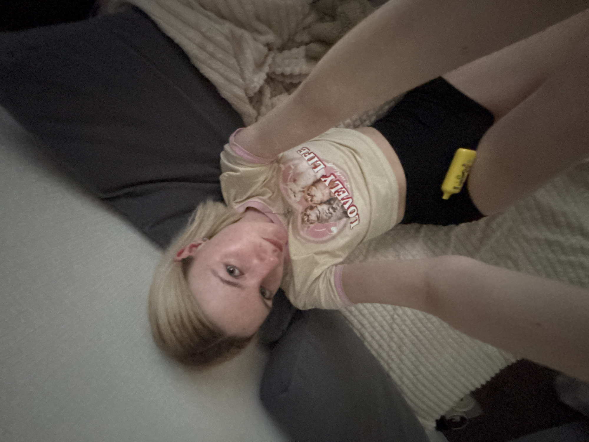 A blonde-haired individual lies on a bed with grey and beige textured surfaces. They are wearing a t-shirt with the words "LOVELY LIFE" printed on it, as well as black shorts. A yellow object is tucked into the waistband of the shorts. The location is Unknown, Unknown.