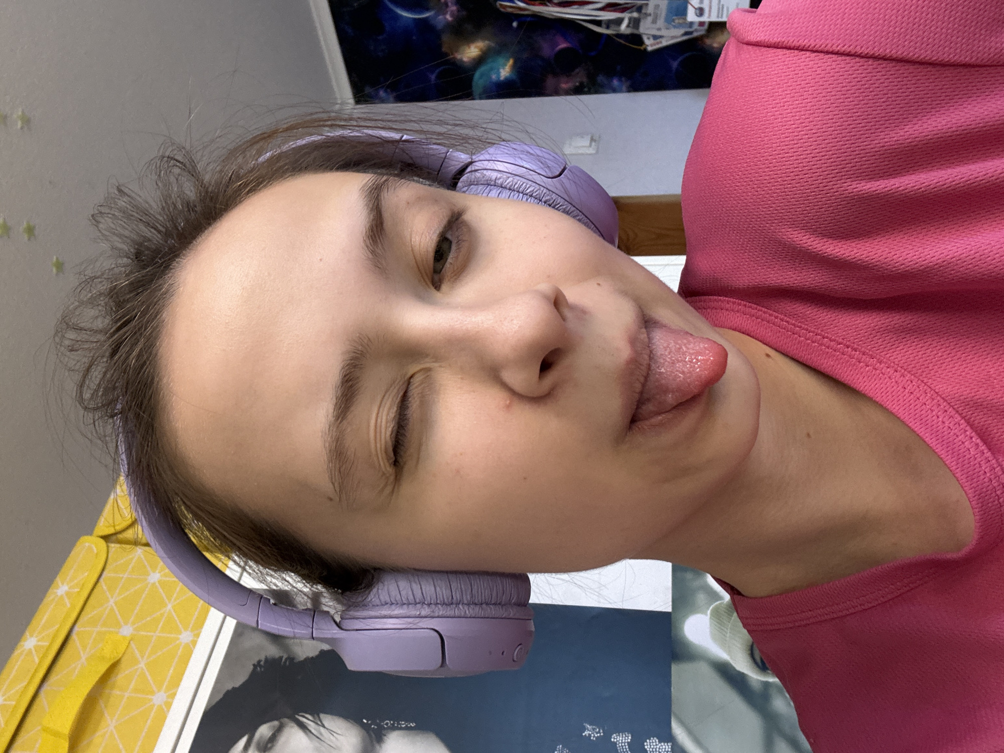 A young woman is pictured in an unconventional, close-up shot. She has brown hair and is wearing light purple headphones. She is wearing a pink top. Her face is the prominent feature, with her left eye closed in a wink and her tongue sticking out. The location cannot be identified based on visual information.