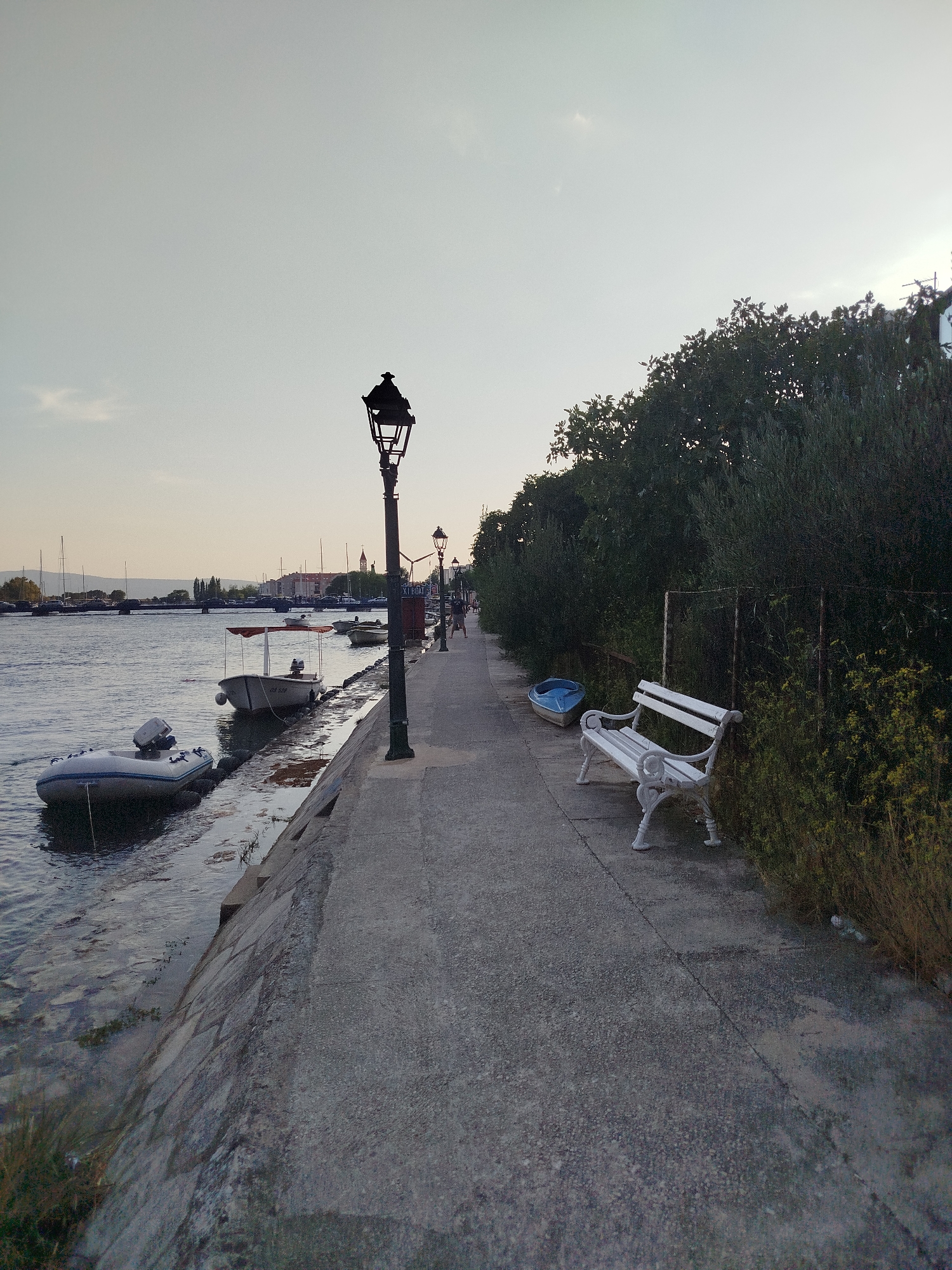 The image depicts a waterfront promenade in Omiš, Croatia. The promenade is paved with concrete and runs alongside a calm body of water, likely the Adriatic Sea. Several boats are moored along the edge of the water, including an inflatable dinghy with an outboard motor and a small white boat with a canopy.
To the right of the promenade, a dense line of trees and shrubs creates a green buffer. A white ornate park bench sits prominently on the promenade, facing the water, inviting repose. Further down the promenade, another, smaller lamppost is visible. In the distance, a town skyline with church spires can be seen across the water, characteristic of Omiš's historic architecture.
The sky is overcast with a pale, diffused light, suggesting it might be late afternoon or early morning. The overall atmosphere is serene and quiet, with no active human interaction or discernible actions taking place in the immediate foreground. The scene captures a typical coastal town vista, highlighting the integration of urban infrastructure with the natural beauty of the waterfront.