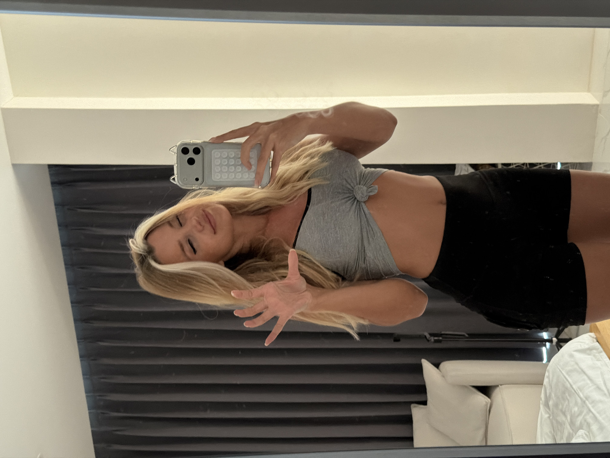 A blonde woman with her eyes closed and a smile takes a mirror selfie in what appears to be a hotel room in Thep Krasatti, Thailand. She is wearing a grey sports bra tied at the front and black athletic shorts. Her left arm is raised, with her hand open and fingers spread. The background shows a dark curtain, a white couch with pillows, and a bed with white linens. A white and grey phone case with cat ears is visible on her phone.