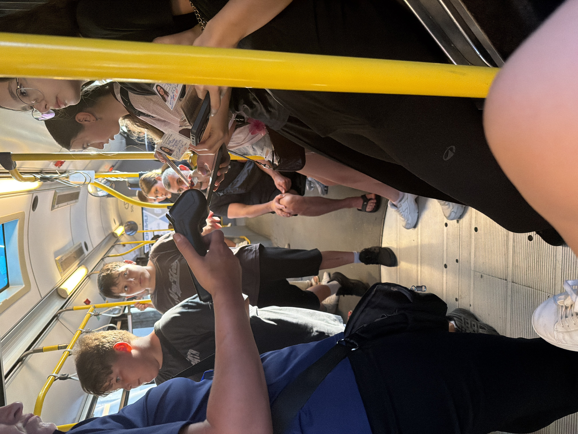 The image captures the interior of a crowded public bus, likely in Burgas, Bulgaria, as indicated by the presence of what appear to be Bulgarian IDs or passes held by passengers. The scene is dominated by yellow poles and handrails, typical of public transportation. Several individuals are visible, primarily young people and adults, standing and seated.
In the foreground, a person wearing a blue shirt is partially visible on the left, holding a black wallet or small bag. To their right, a young boy with brown hair looks to the left, and another boy with dark hair is looking forward. Further back, a woman with glasses and a light-colored top is holding a phone, and a woman with dark hair is seated. A man in a dark t-shirt and shorts is seated in the middle ground. The floor of the bus is a light-colored ribbed material, and overhead lights and windows are visible. Some passengers are holding what appear to be identification cards or transit passes. The overall impression is of a busy, mundane commute.