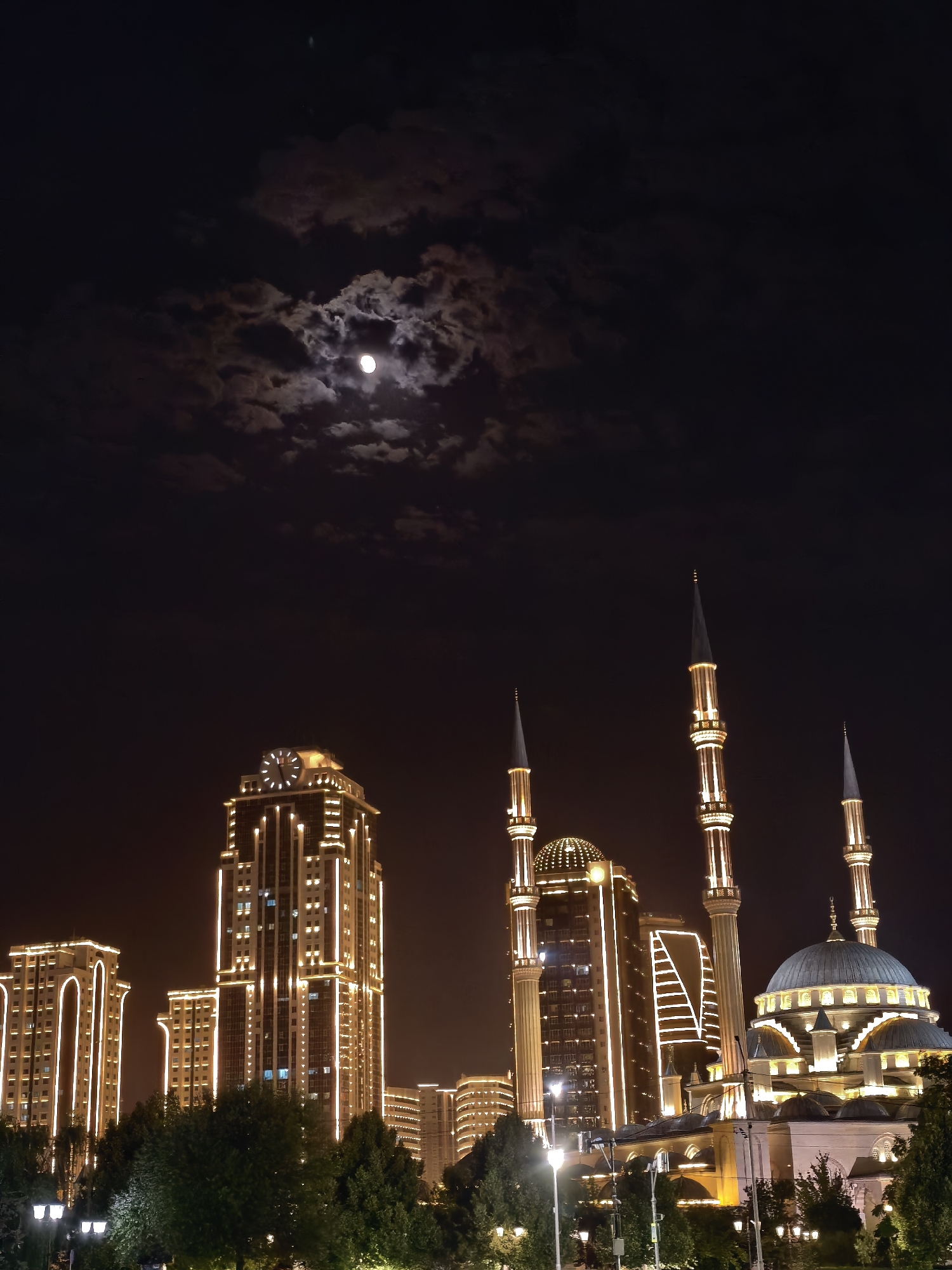 A night view of the city of Grozny, Russia, showcases a prominent mosque illuminated by lights, with several modern high-rise buildings visible in the background. The moon is partially obscured by clouds, casting a soft glow on the scene. The mosque features a large dome and four tall minarets, all adorned with lights. The buildings in the background are also brightly lit, highlighting their architectural details. Trees and streetlights are visible in the foreground, suggesting a park or public area. The overall atmosphere is serene and majestic.