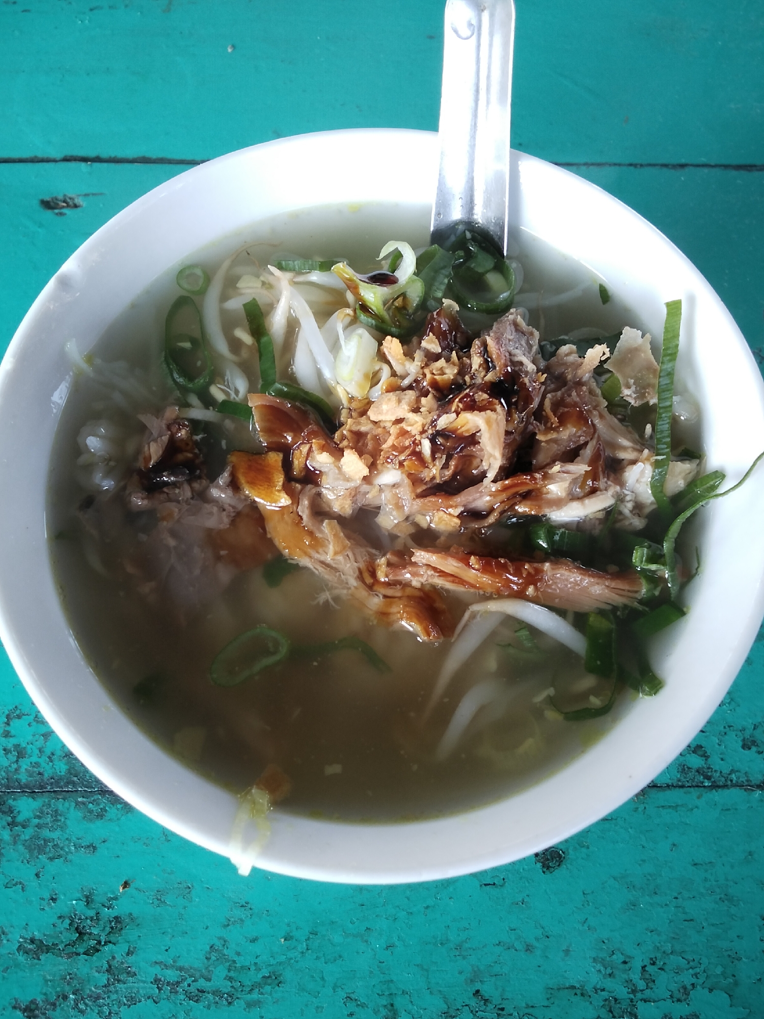 A white bowl filled with a hearty broth, shredded chicken, bean sprouts, and green onions rests on a teal, distressed wooden surface. A silver spoon is partially submerged in the broth, its handle extending upwards and out of the frame. The lighting suggests an outdoor or well-lit indoor setting. The image appears to be taken at eye level, looking directly down into the bowl. The overall impression is a close-up view of a delicious and freshly prepared meal.
