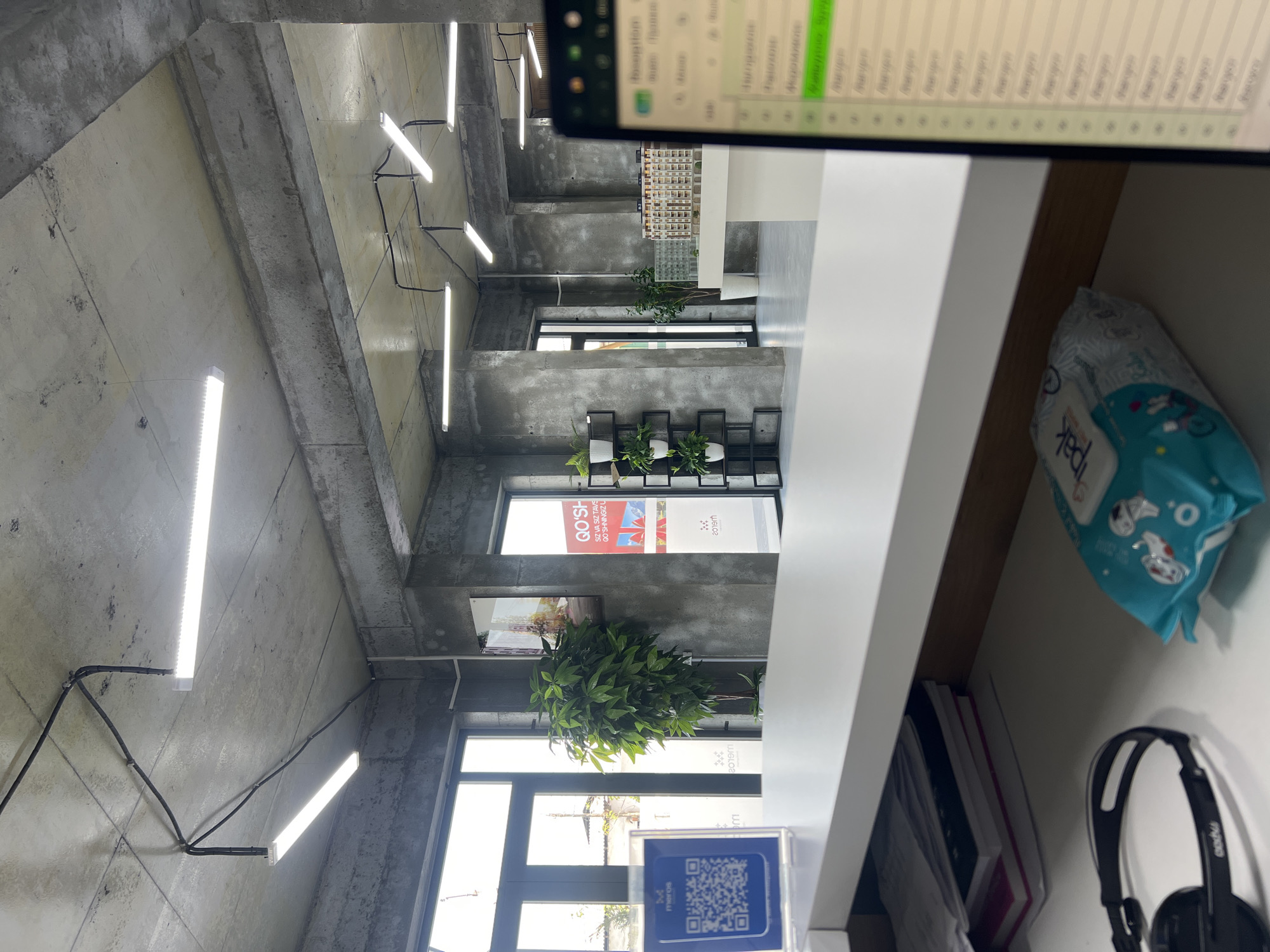 The image depicts the interior of a modern office space, characterized by exposed concrete walls and ceilings, typical of an industrial or loft aesthetic. Linear LED lights are installed on the ceiling, providing illumination. The environment appears to be a reception area or an open-plan workspace.
In the foreground, a white reception desk is visible, with a stack of books and a pair of black headphones on the surface. To the right, the edge of a laptop screen shows a list of items, possibly a contact list or a system interface. A blue QR code displayed in a holder is also present on the desk.
Further into the space, a tall potted plant with lush green leaves stands near a large window. Beyond that, a miniature architectural model is displayed on a white surface. The background reveals doorways leading to other areas of the office, one of which has a sign with text that appears to be in Uzbek, suggesting the location is in Uzbekistan. Based on the Uzbek text and the visual cues of a modern office environment, it is plausible this scene is located in the city of Shahrixon, Uzbekistan. The overall impression is one of a contemporary, clean, and functional workspace.