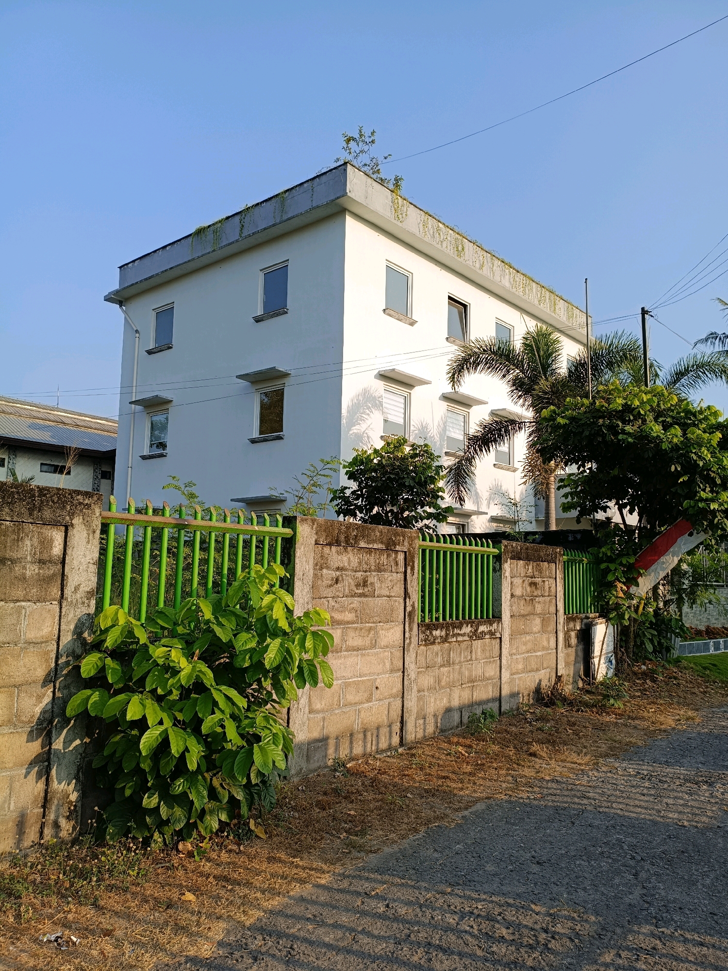 The image depicts a two-story white building with several windows on each level, located in Maguwoharjo, Indonesia. The building has a flat roof with some vegetation growing on it. In front of the building, there is a concrete block wall topped with a green metal fence. Lush green bushes grow in front of the wall. A paved road runs alongside the fence, and shadows from the fence and vegetation are cast upon it. Palm trees and other trees are visible to the right of the building. The sky is clear and blue.