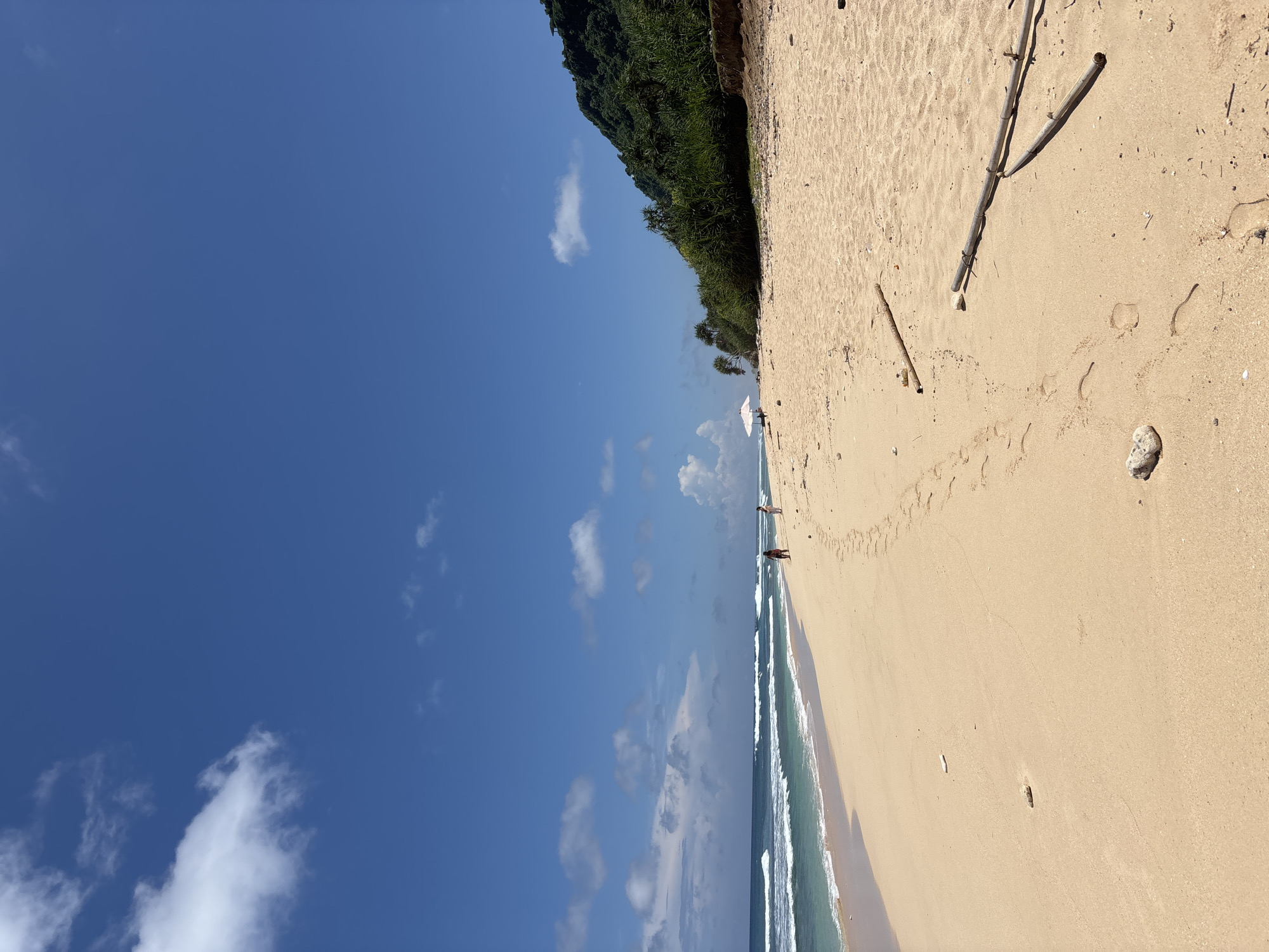 A beach in Pecatu, Indonesia, shows a blue sky with scattered clouds, the ocean with small waves, and a sandy shore. Lush green vegetation borders the beach. Two people stand near the water's edge. The sand is marked with footprints and scattered debris like driftwood and a rock.