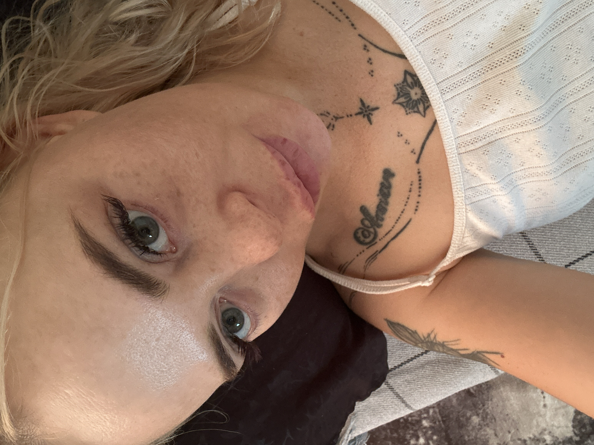A close-up, top-down selfie of a young Caucasian woman with blonde hair and blue eyes is shown. She is wearing a white, sleeveless, ribbed top with a delicate eyelet pattern. She has several tattoos visible on her chest and upper arm. One tattoo on her chest reads "Amour" in cursive script, with floral and star designs surrounding it. Another tattoo on her upper arm appears to be of a winged creature or insect. The woman's skin is light-toned and shows some freckles and minor blemishes. Her eyebrows are dark and well-defined, and she is wearing mascara on her eyelashes. Her lips are painted a light pink. She is lying down, with her head tilted and resting on a dark surface that is partially visible. A grey and white checkered fabric is also visible beneath her arm. The background is indistinct, suggesting an indoor setting. The lighting is bright, casting some shadows on her face.