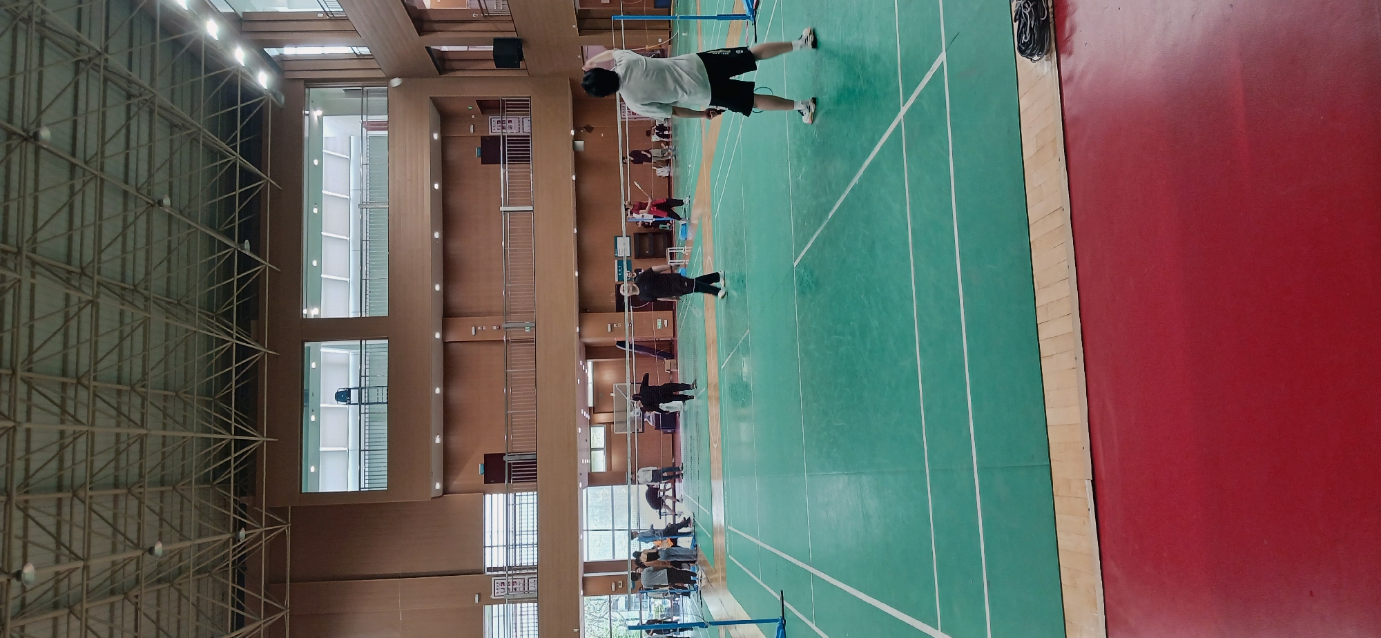 The image captures an indoor badminton court within a sports facility, likely located in the Chongqing High-tech Zone, China. The environment is well-lit by overhead fluorescent lights, illuminating a green badminton court delineated by white lines. The court is bordered on one side by a red-surfaced area. Players are actively engaged in badminton, with one individual in mid-swing, poised to hit the shuttlecock. Spectators are seen observing the game along one side of the court. The sports facility's interior features a tall ceiling supported by a steel structure, and walls clad in wood paneling with windows providing natural light.