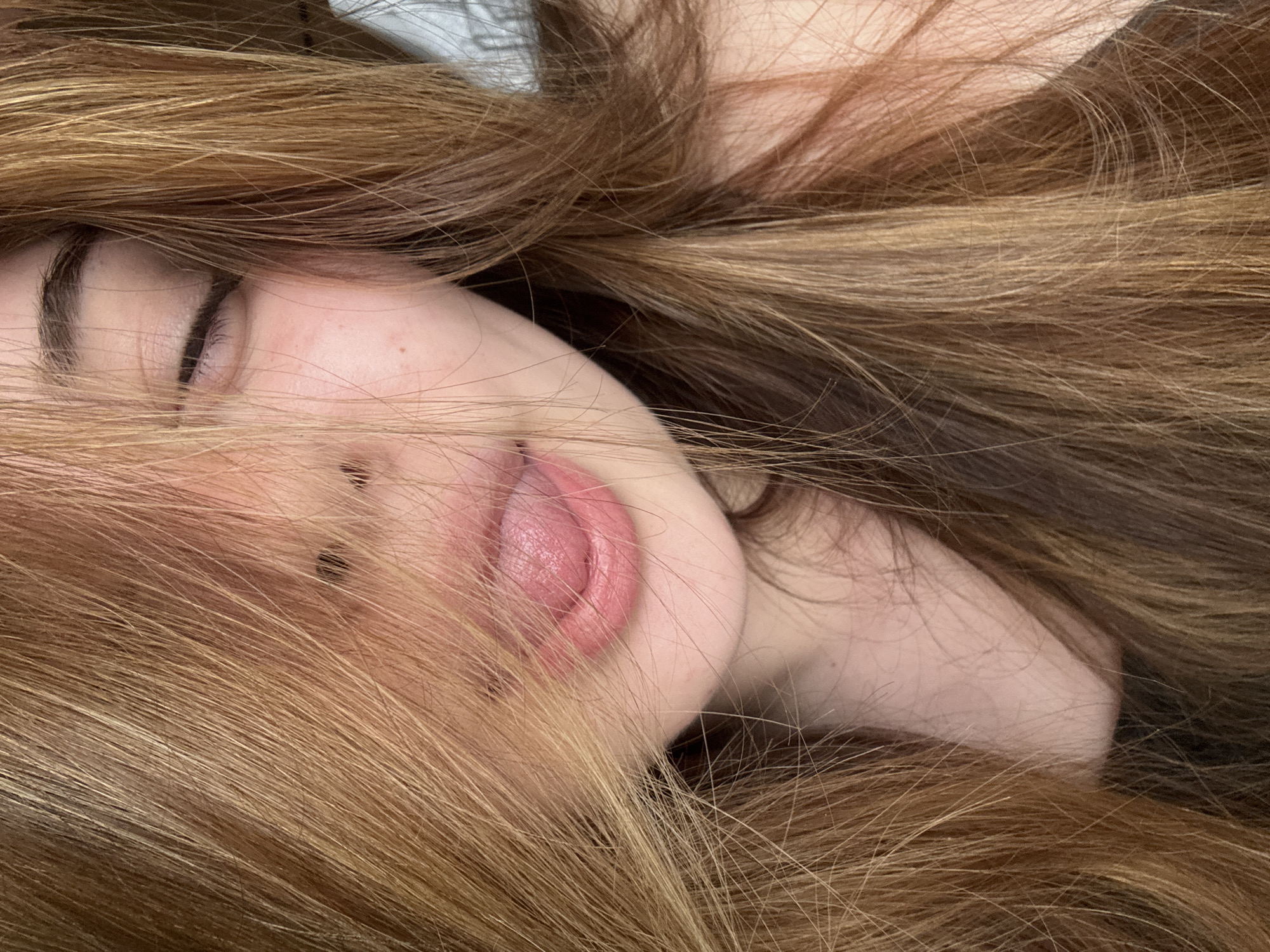 A close-up, intimate portrait captures a person lying down, with their long, flowing hair framing and partially obscuring their face. The dominant feature is a mass of multi-toned light brown, golden, and auburn hair, appearing thick and slightly wavy, cascading across the frame and around the subject's head.
The visible parts of the face include a closed eye with a defined eyebrow and delicate eyelashes, the bridge of the nose, and the mouth with slightly parted pink lips and the tongue subtly extended. The skin appears fair, with a few faint freckles or beauty marks visible on the cheek. The person's expression, with the closed eye, suggests a state of relaxation, sleep, or a dreamy contemplation. The partially extended tongue adds a hint of playfulness or sensuality.
The composition is very tight, focusing solely on the subject's head and hair, creating a sense of intimacy and soft enclosure. There are no discernible background elements, location cues within the image, visible objects beyond the person, or text. While the location context is provided as Troyan, Bulgaria, this information is external to the visual content of the image.