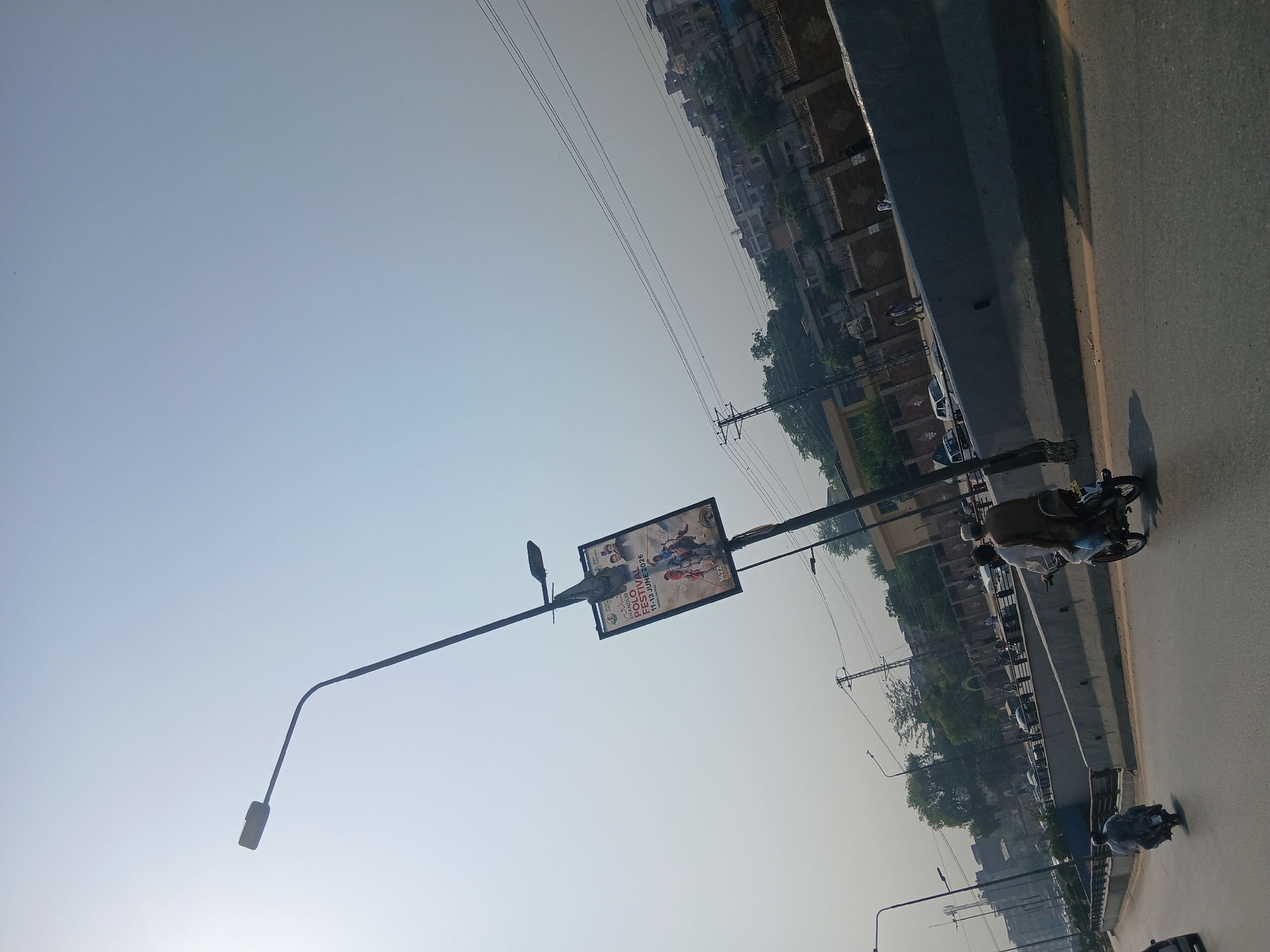 The image captures a street scene in Peshawar City Tehsil, Pakistan, on a clear day. A large street lamp with a prominent sign attached dominates the center-left of the frame. The sign advertises a "POLO FESTIVAL" scheduled for "11-13 JUNE 2026". The festival appears to be supported by "Jhandur" and possibly a radio station. In the foreground, a road stretches out with several individuals on motorcycles. One motorcycle, in the lower right, carries two men; the driver wears a light brown shalwar kameez, and the passenger wears a darker brown shalwar kameez. Another motorcycle is visible further down the road to the left, with a single rider. The road is flanked by concrete barriers and a slight incline on the right side, with buildings and trees visible in the background, suggesting an urban or semi-urban environment. Power lines and a utility pole are also present, indicating infrastructure in the area. The sky is a pale blue and hazy, possibly due to atmospheric conditions or dust. The overall impression is that of a busy thoroughfare in a Pakistani city.