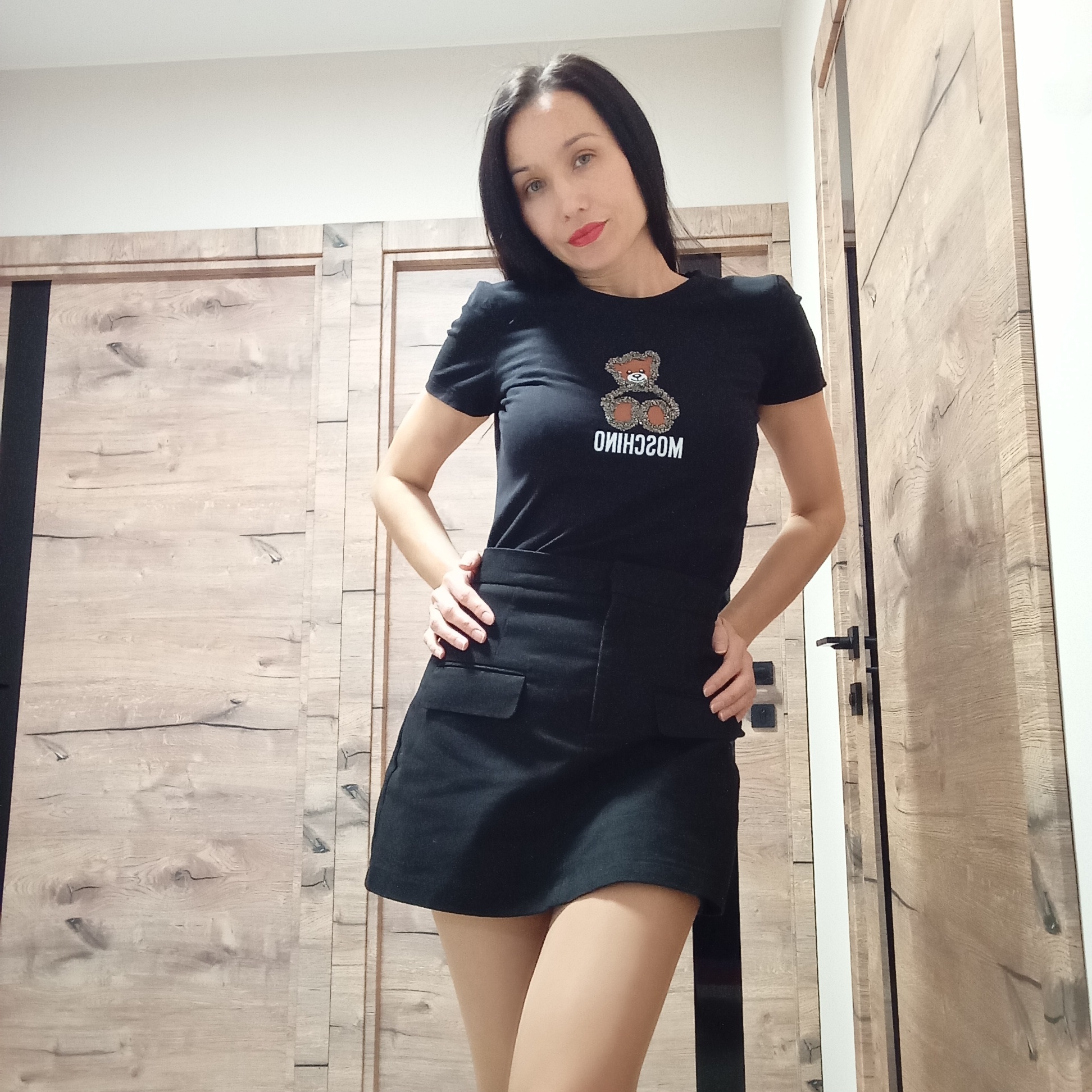 A woman with long, dark hair stands indoors in front of two wooden doors. She wears a black T-shirt with a graphic of a teddy bear and the word "MOSCHINO" printed upside down below it. She is also wearing a black skirt and flesh-colored tights. She has her hands on her hips and is looking at the camera. The location appears to be an interior space with white walls and wooden doors. There is no indication of this being Pervomaysky, Russia.