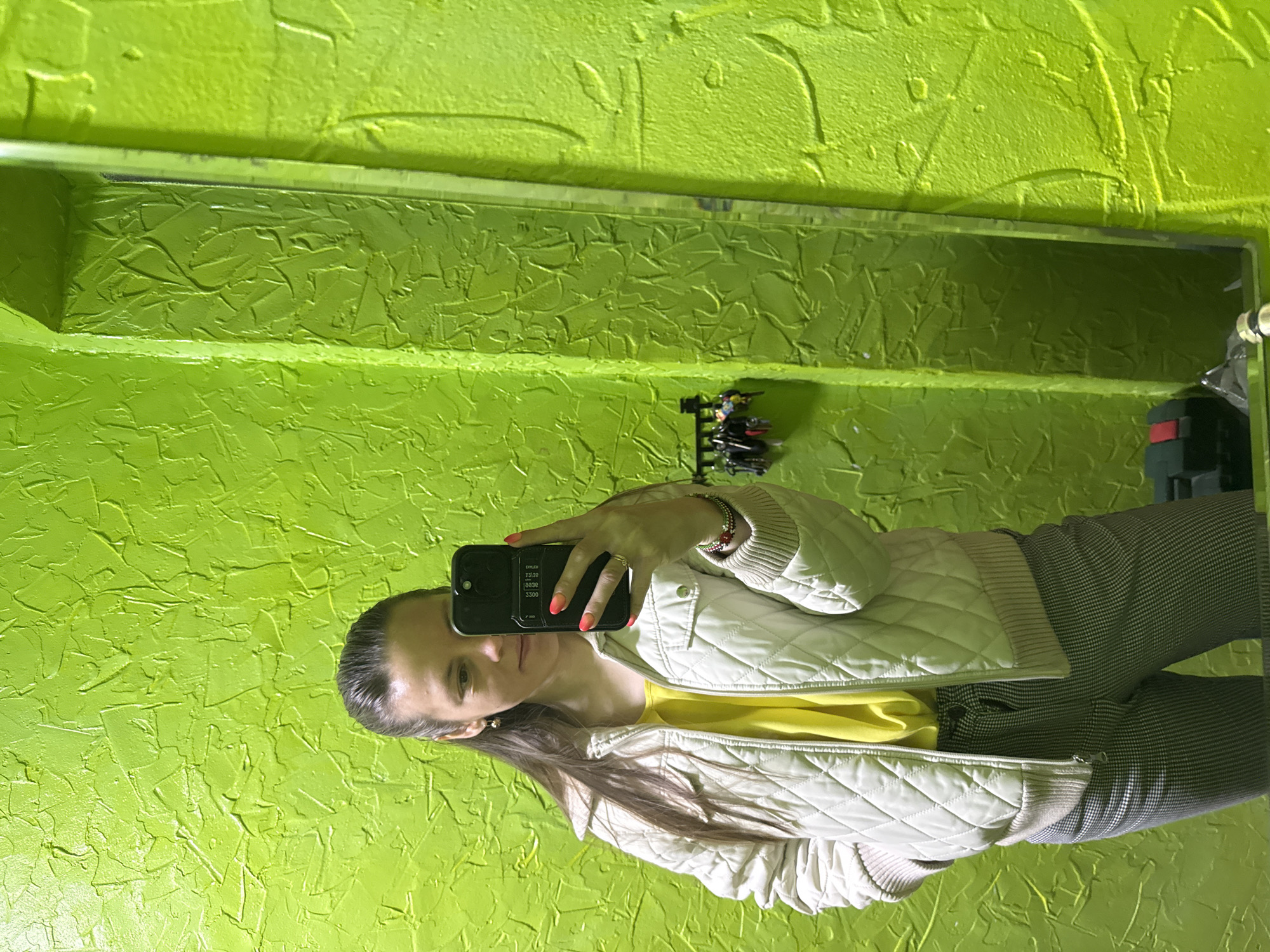 In an interior setting in Chaikovsky, Russia, a woman takes a mirror selfie with her phone. The image is rotated 90 degrees clockwise, showing the woman positioned vertically along the left side of the frame, holding a black smartphone. She is reflected in a mirror against a vibrant, textured lime green wall, which features a distinct, uneven plaster-like finish.
The woman has long dark hair pulled back and is wearing a light beige quilted bomber jacket over a bright yellow t-shirt. Her lower body is partially visible, showing dark and light plaid trousers. On her right wrist, she wears several bracelets, including some beaded and some metallic. A ring is visible on her right hand, and her fingernails are painted with a bright orange-to-pink gradient polish.
The black smartphone has a case with visible card slots on the back. Due to the mirror reflection, text on the phone screen appears reversed, showing what looks like "2T.J.3P," "0030," and "3300."
Behind her reflection, the textured green wall continues, with a narrow silver-colored shelf or trim running horizontally across the mid-section of the wall. Below this, another shelf holds a small rack of keys and some other indistinct objects. The overall impression is a casual, personal photo taken in a distinctively decorated space.