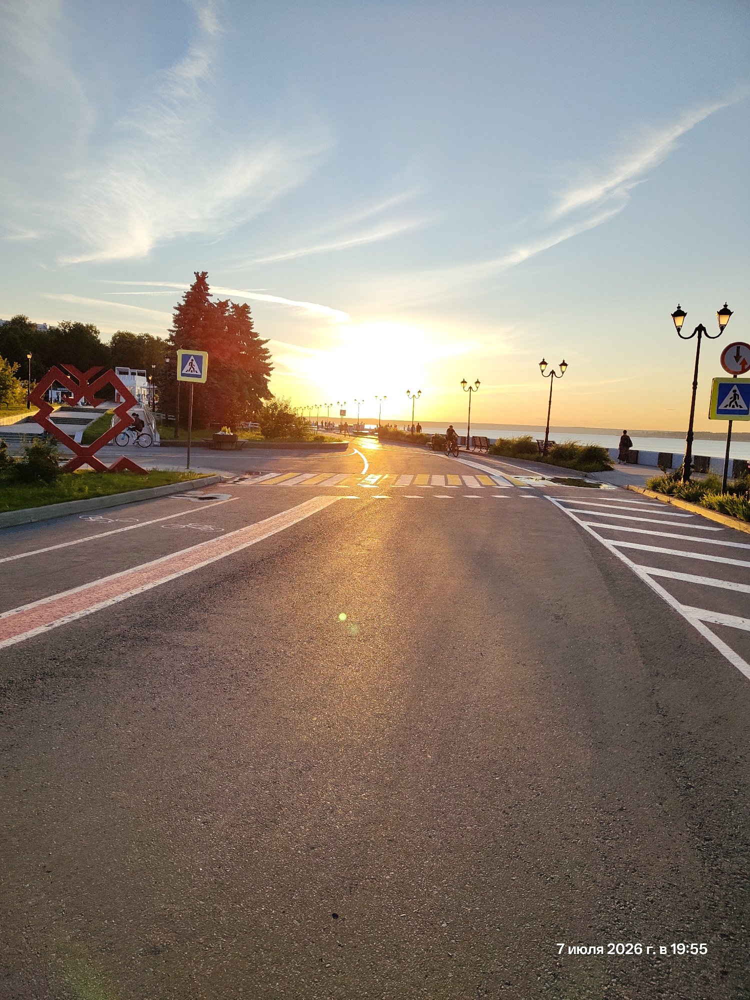 The image depicts a street scene at sunset in Unknown, Unknown. The sun is low on the horizon, casting a warm, golden light across the asphalt road, which is marked with white lines for a pedestrian crossing and a bicycle lane. To the left of the road, a large, red, abstract heart-shaped sculpture is visible, along with some trees and a building. Further down the road, streetlights illuminate a walkway beside a body of water, with several people walking and cycling. The sky is clear blue with wispy clouds, and the overall atmosphere is serene and peaceful. A timestamp at the bottom right corner indicates "7 июля 2026 г. в 19:55" which translates to "July 7, 2026 at 19:55".