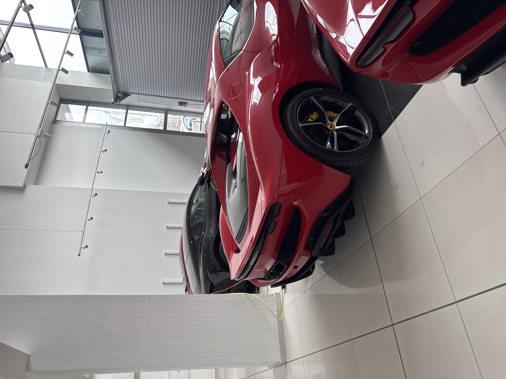 The image shows the front ends of two luxury cars parked in a showroom. The car in the foreground is red, and the one behind it is black. The red car's wheel has a yellow Ferrari logo. The floor is tiled in white. There are structural elements in the foreground and the background of the image, likely elements of the showroom's architecture. The location is Sofia, Bulgaria.