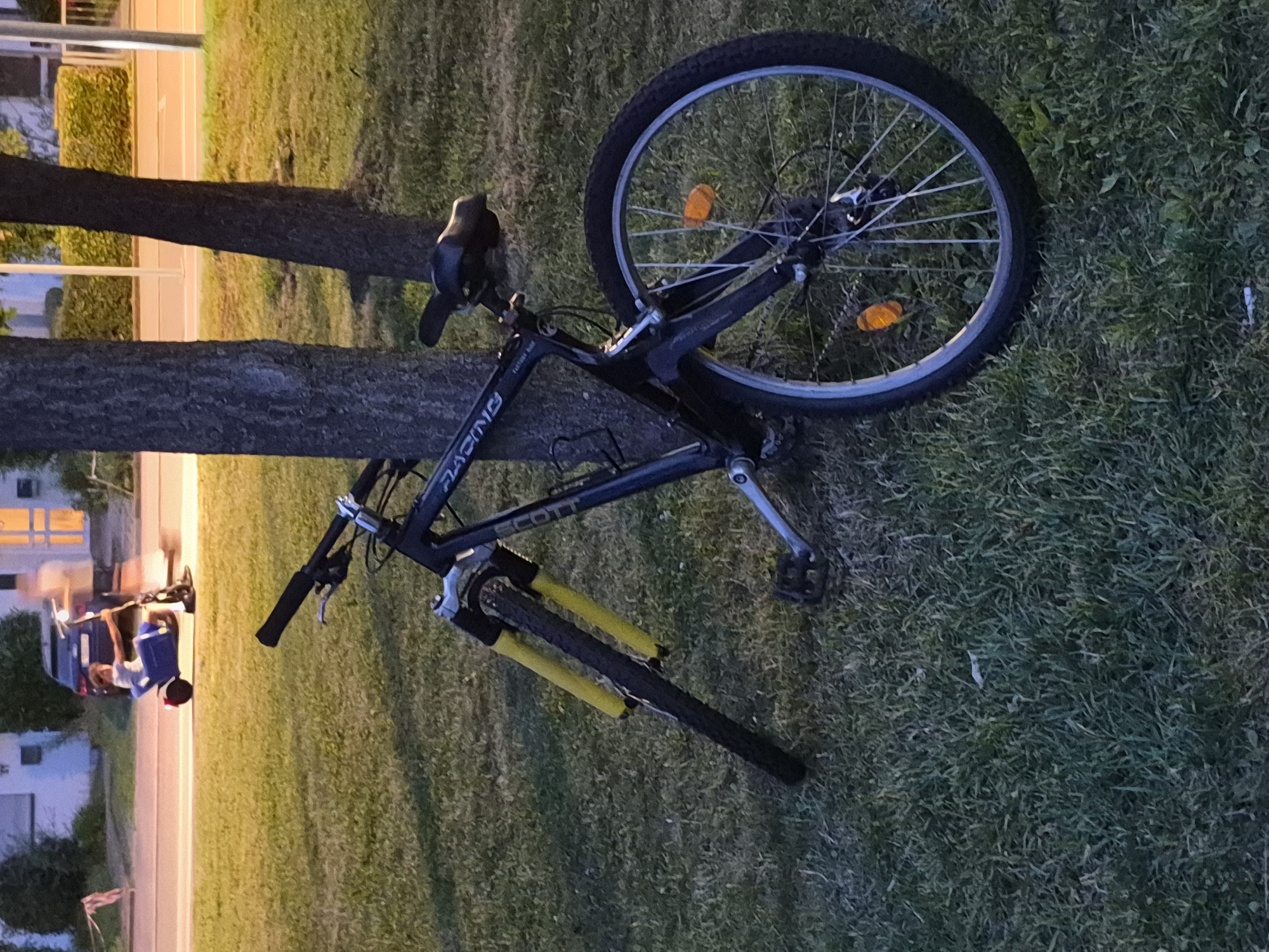 An outdoor scene at dusk features a dark blue mountain bike leaning against a tree trunk on a grassy area. The bike has yellow front suspension forks, a black seat, and a black frame with "RACING" written on it. The front wheel is angled, and the handlebars are turned left. In the background, across a paved area, a person is partially visible driving a blue motorized scooter with a person riding on the back. Buildings and trees are also visible in the background. The lighting suggests artificial light sources are present, casting a warm glow.