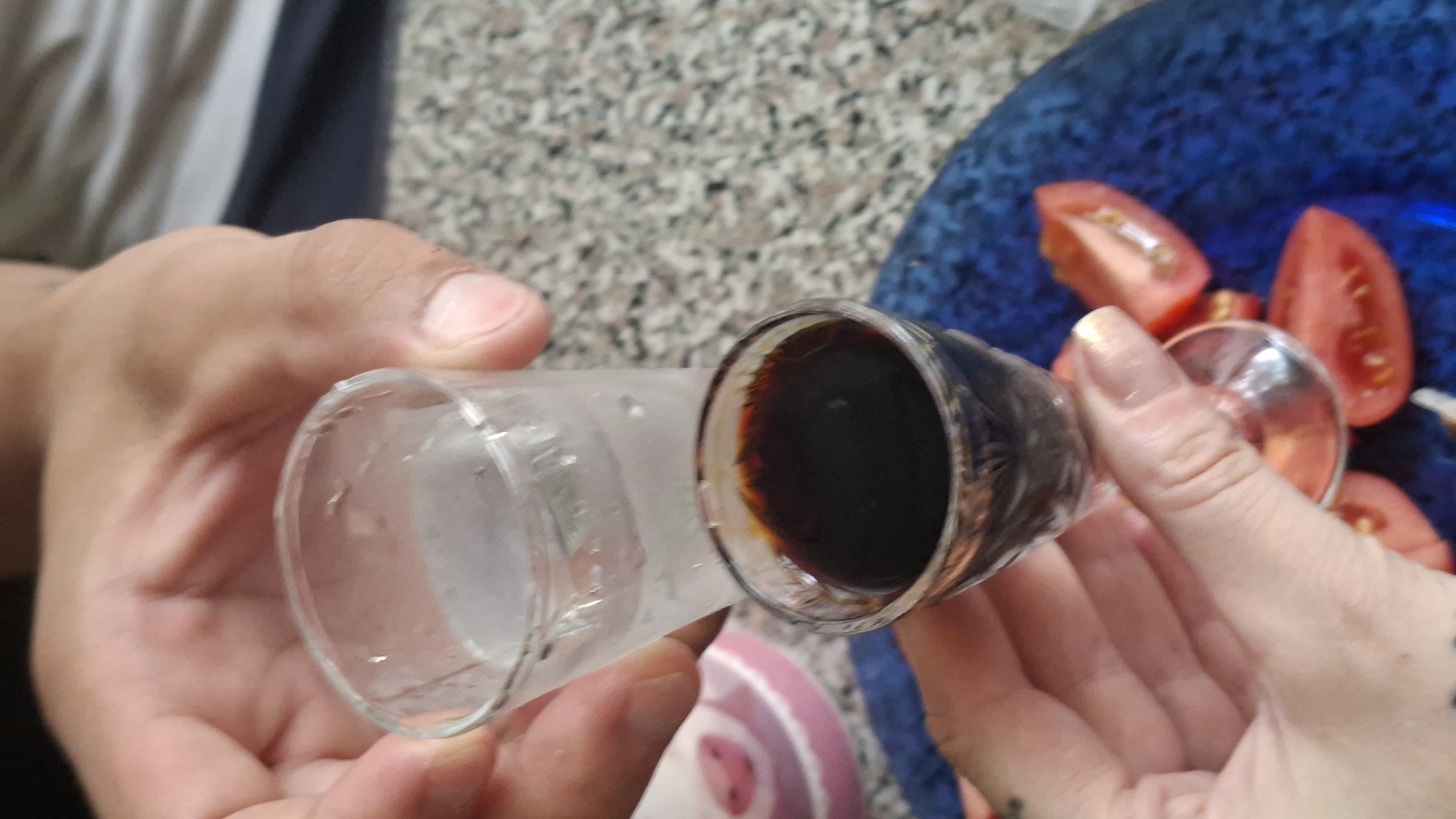 A close-up shot shows two hands holding small shot glasses. The glass in the foreground contains a dark liquid, possibly liqueur or a dark colored beverage, and is being held by a hand with manicured fingernails. Behind it, another hand holds a glass containing a lighter, translucent liquid, likely water or vodka. The background suggests an indoor setting with a speckled countertop and a blue plate with sliced tomatoes. The context of Myadzyel, Belarus is not visually discernible from the image itself.