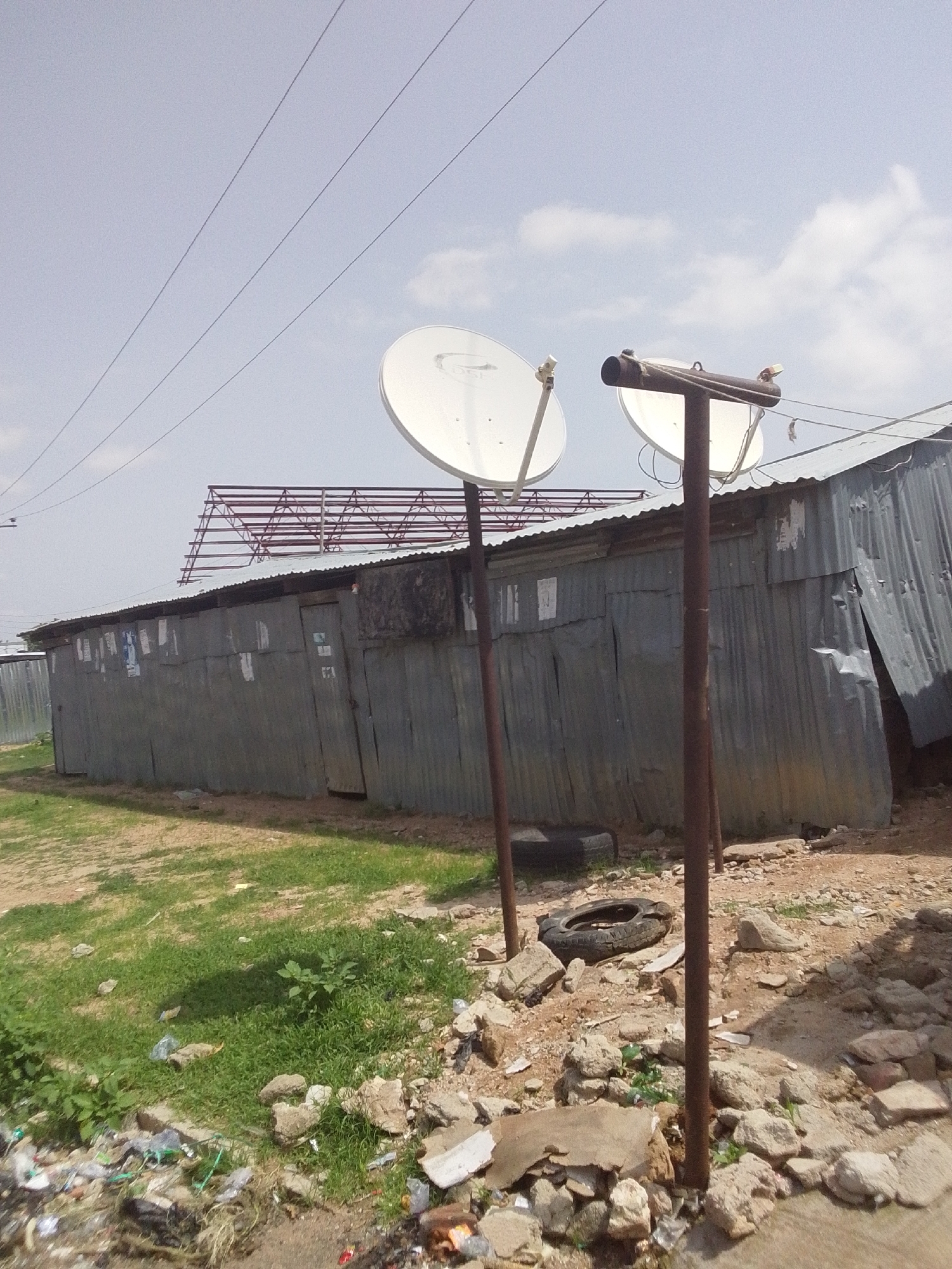 In Jalingo, Nigeria, this outdoor scene captures a cluster of corrugated metal structures, likely makeshift dwellings or workshops. Two satellite dishes are prominently mounted on poles in front of one of the structures, suggesting a need for external communication or entertainment. The surrounding environment is one of disarray; the ground is uneven and littered with debris, including scattered trash, rocks, and a discarded tire. Vegetation, primarily grass, is sparsely growing amidst the rubble, indicating a neglected or undeveloped area. Overhead, power lines traverse the sky, and a skeletal metal frame, possibly an unfinished structure or advertisement billboard, is visible in the background. The overall impression is one of a struggling urban environment where basic infrastructure coexists with visible signs of poverty and disrepair.
