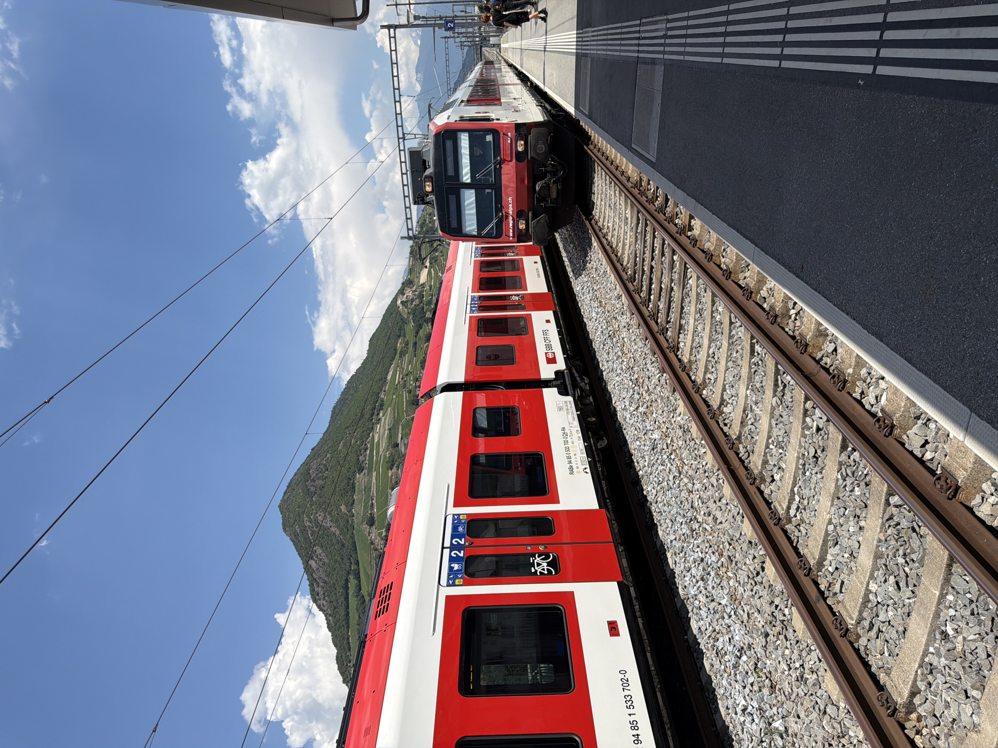 An eye-level, vertical, medium shot captures two SBB (Swiss Federal Railways) trains at the Saint-Léonard train station in Switzerland on a bright, sunny day. The train on the left is predominantly white with a red stripe along the top and a red section around the doors and windows. It is stopped at the station, and a number "2" is visible on the side of the train, above a set of red doors with icons for luggage, a person, and a bicycle. Below the window, the number "94 85 1 533 702-0" is printed. The train on the right is also primarily white and red, but its front is more visible, featuring a red nose cone with headlights. This train also has "www.regional.ch" printed on its side. It appears to be either arriving or departing from the station, positioned on a parallel track.
The tracks themselves are clearly visible in the foreground, laid on a bed of gravel with concrete sleepers. To the right of the tracks is the station platform, a dark asphalt surface with tactile paving strips indicated by parallel white lines. In the background, a lush green mountain looms, with terraced vineyards visible on its slopes, suggesting the location is in a wine-growing region. The sky is a vibrant blue, dotted with fluffy white clouds. A few people are visible on the platform in the distance, near the train on the right, dressed casually. The overall impression is a peaceful, operational scene at a Swiss railway station, set against a scenic mountainous backdrop.