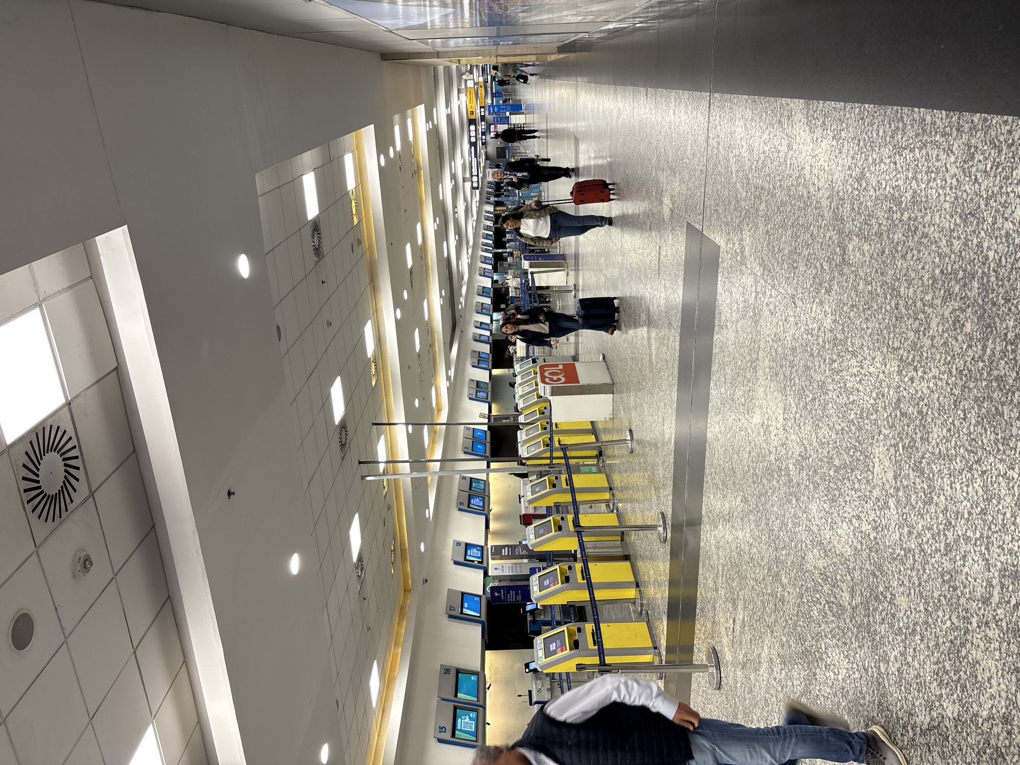 This image depicts an indoor airport terminal check-in area at Aeroparque Jorge Newbery (AEP) in Buenos Aires, Argentina. The environment features a bright, spacious hall with a white false ceiling composed of rectangular panels, square recessed fluorescent lights, and circular ventilation grilles. The floor is made of light gray speckled terrazzo or granite tiles, with a darker gray section running along the right wall.
On the left side of the scene, a long row of check-in counters is visible, each identified by a blue display screen with a white number, ranging from 15 to at least 20. In front of these counters, a parallel line of approximately 15-20 yellow self-check-in kiosks is arranged, separated by silver stanchions and blue barrier tapes. Signs for "LATAM" are present above some kiosks, and a red and white "GOL" branded counter is in the left foreground. The right wall displays large, framed advertisements or informational posters. Further in the background, airport signage, including "Control de Seguridad" (Security Control), is discernible.
Approximately 10-15 individuals are present, primarily in motion. In the immediate foreground, a man in a dark blue quilted vest, light blue shirt, and jeans walks from left to right. In the mid-ground, two women walk towards the viewer: one in a dark jacket and jeans pulls a dark blue roller suitcase, while the other, also in a dark jacket and jeans, pulls a red roller suitcase. Behind them, a man in a suit jacket walks away from the viewer, holding a bottle. Other individuals are visible in the distance, walking with luggage. People are engaged in typical airport transit, moving through the check-in area towards other sections of the terminal. No direct interactions between individuals are clearly evident.