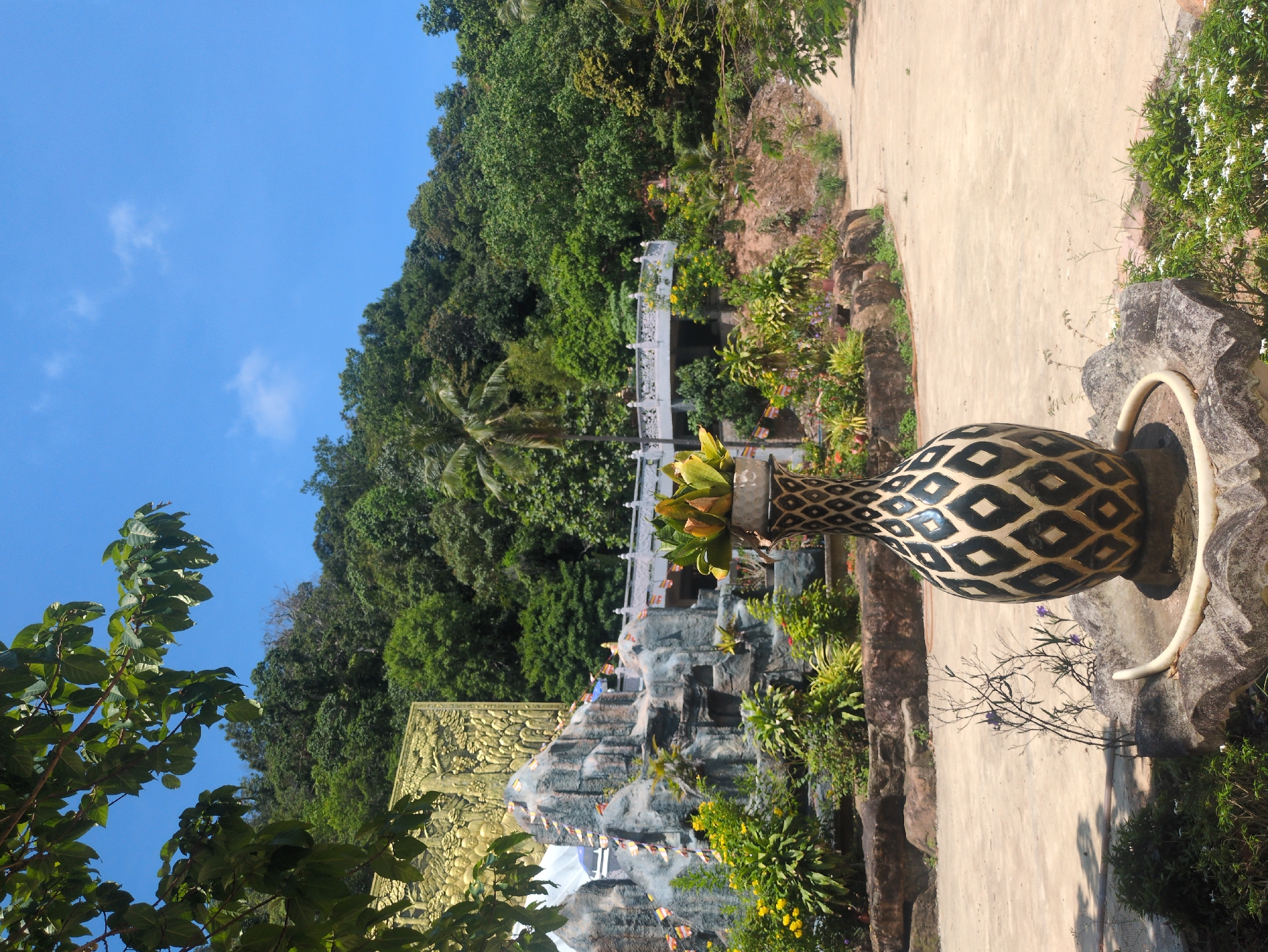 The image depicts a landscape at a temple in Phú Quốc, Vietnam. In the foreground is a large decorative vase or urn with a diamond pattern, positioned along a paved walkway. To the left and beyond the vase, a sculpted golden bas-relief artwork can be seen at the foot of a man-made waterfall with figures. A bridge is located in the background amidst lush green trees. The sky is a clear blue with a few wisps of clouds.