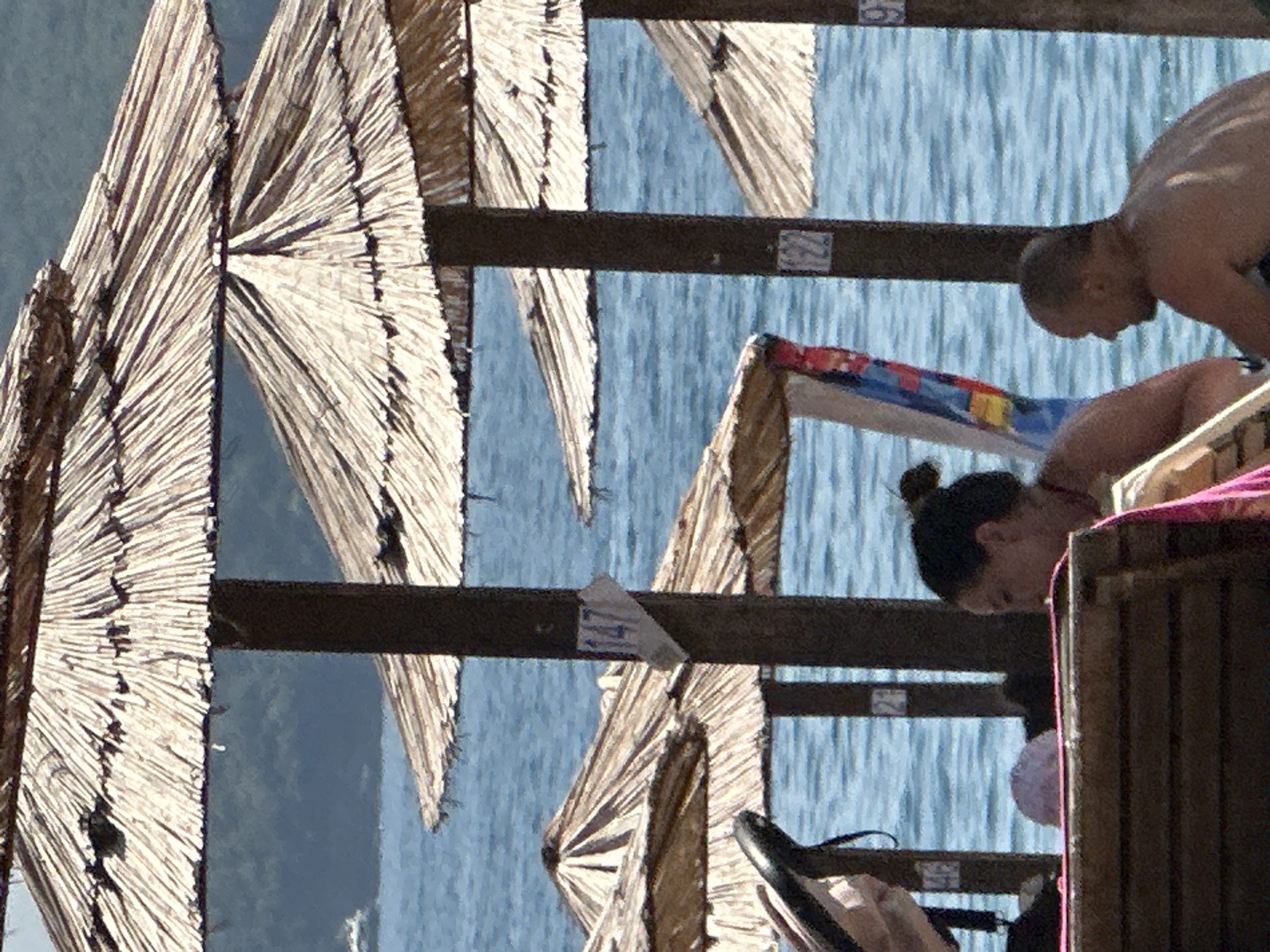 The image captures a sunny day at a beach in Budva, Montenegro, with a focus on straw umbrellas and the sea. In the foreground, a wooden beach chair is partially visible, with a person's back facing the camera, wearing a pink towel draped over their shoulder. To the right, a man with a shaved head is seated, looking out towards the sea. Behind them, a woman with her hair in a bun is also seated, looking towards the man. The sea is a vibrant blue, with gentle waves. Several straw umbrellas are positioned on the beach, providing shade. A sign with the number "147" is attached to one of the umbrella poles, indicating a numbered section of the beach. The background shows a hazy coastline with greenery, suggesting hills or mountains. The overall scene depicts a relaxed day at the beach, with people enjoying the sun and sea.