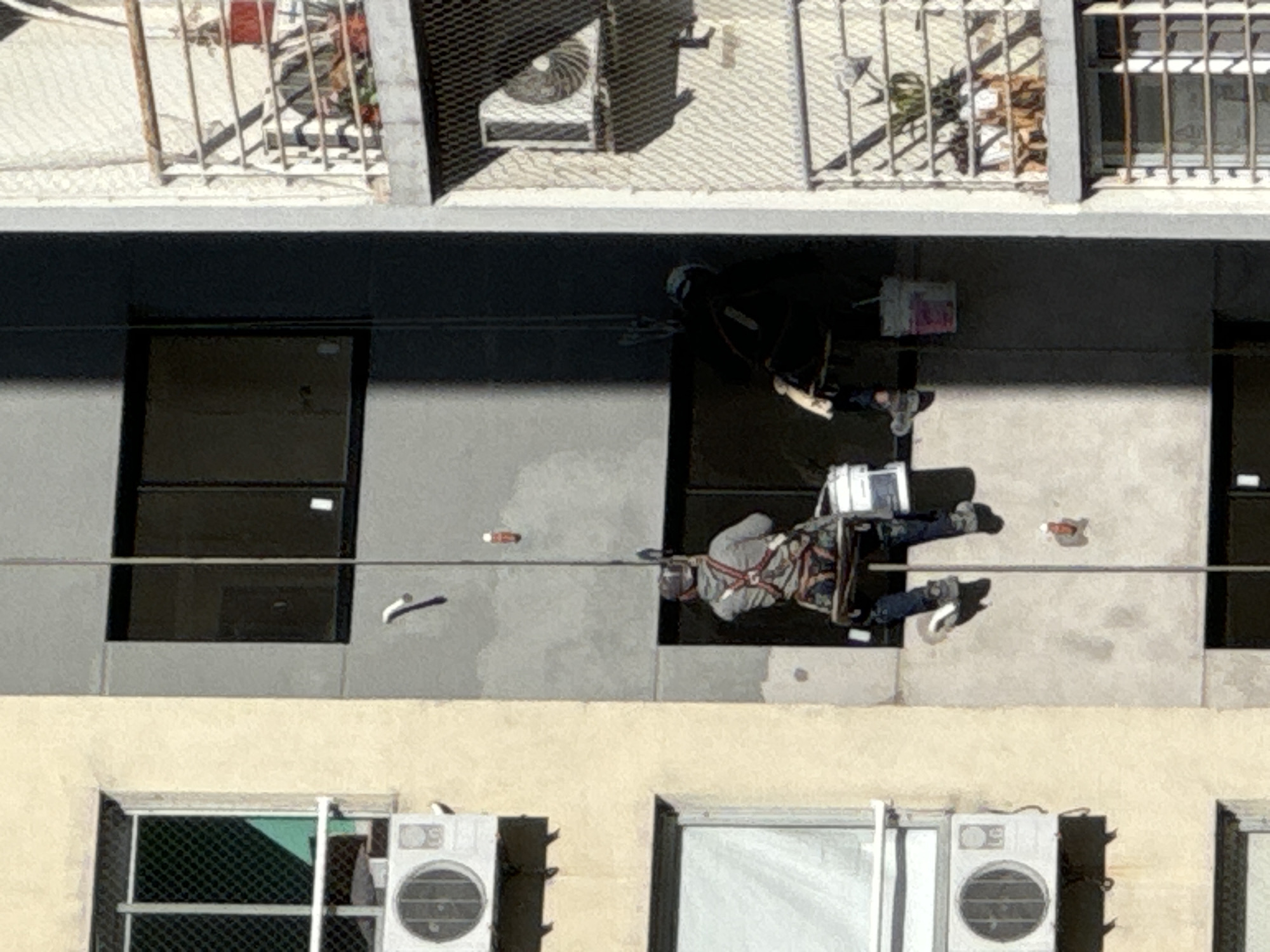 The image captures an exterior view of a multi-story building. Two workers are suspended from ropes performing maintenance on the building's façade. One worker is clad in black, wearing a helmet. The other worker is wearing a grey shirt. They are positioned at different levels of the building. The building has windows and external air conditioning units. There is fencing on the building roof. Based on the architectural style, the building could be located in Buenos Aires, Argentina.