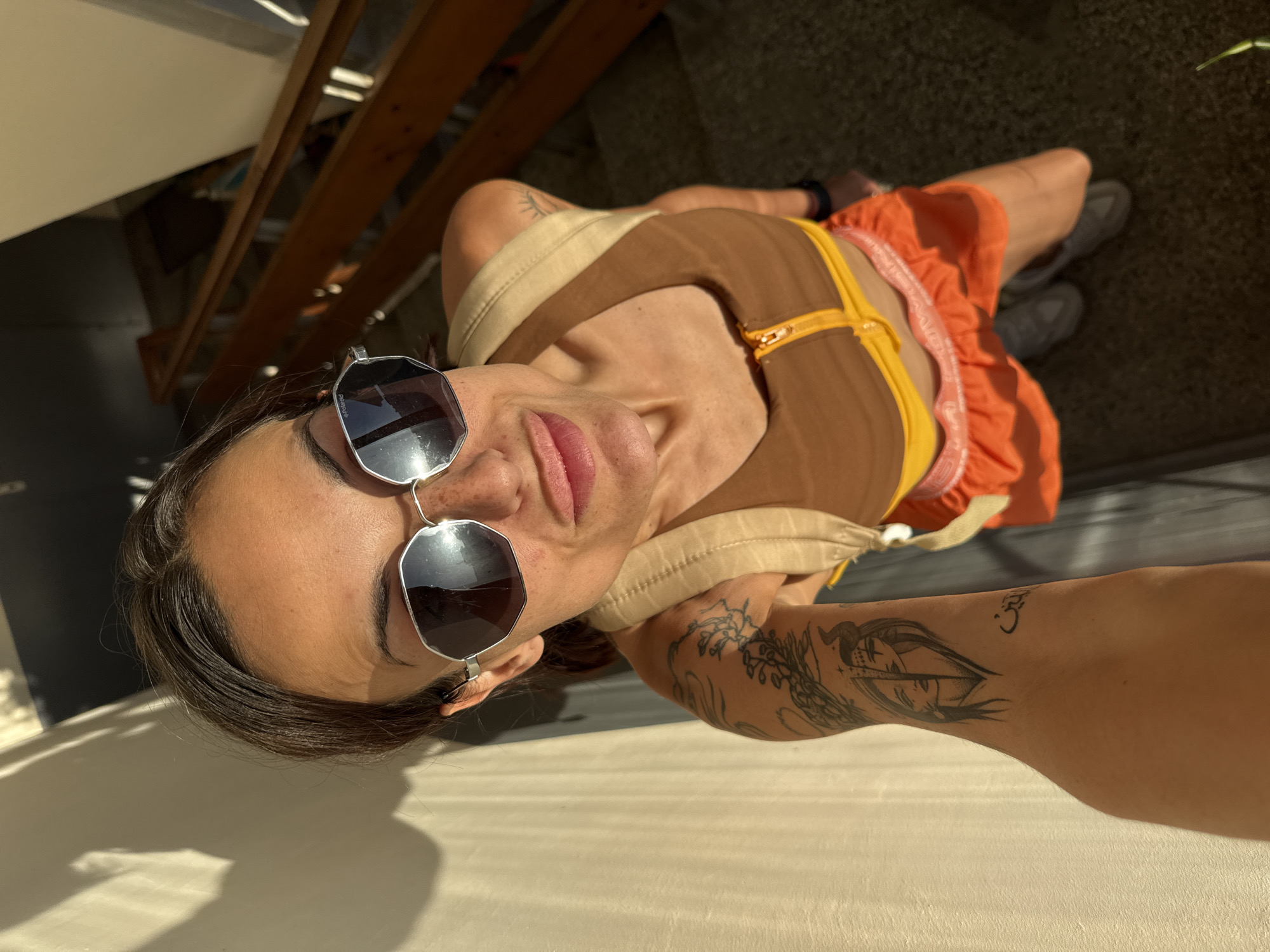 A woman with dark hair, wearing sunglasses, a brown and yellow sports bra, an orange ruffled skirt, and grey sneakers is standing on stairs. Her left arm, which is visible in the foreground, has tattoos, including one that appears to be a split face and another with Arabic script. She is holding a phone to take a selfie. The background shows a wooden railing and a concrete wall, suggesting an outdoor staircase. The lighting indicates it is daytime, with sunlight casting shadows. The image was captured in the city of Pleven, Bulgaria, on a staircase likely located in a residential building or a public space.