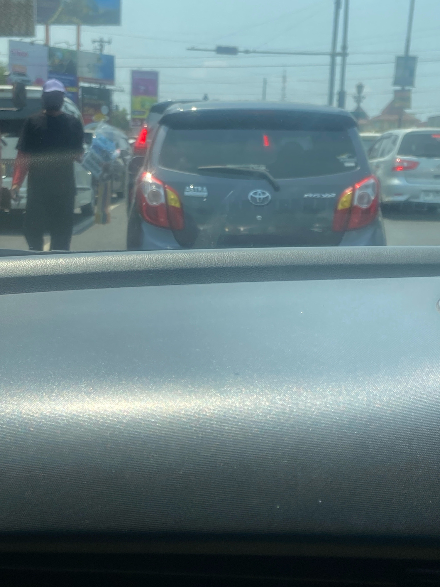 This is a medium shot, eye-level, outdoor photograph taken from inside a vehicle, looking out at traffic. The scene is likely on a street in Ngaglik, Indonesia, as indicated by the presence of a Toyota car with Indonesian license plates, and general signage consistent with the region.
In the foreground, the textured dashboard of the vehicle is visible, reflecting the bright, hazy sky. Beyond the dashboard, a dark gray hatchback car is the most prominent subject. The car is facing away from the camera and is stopped in traffic. Its brake lights are illuminated, a red vertical light is visible on the rear windshield, and the taillights are red and yellow. The Toyota logo is centered on the rear of the car.
To the left of the Toyota, a person in a black t-shirt and a purple cap is walking, carrying what appears to be a bundle of goods. This person is walking in front of another vehicle, partially obscured. To the right of the Toyota, a silver car is also stopped in traffic.
The background shows a hazy sky with overhead wires and what appear to be billboards or signs, typical of a roadside environment in a city. The overall impression is a moment captured in ordinary traffic, likely during daytime, with subdued lighting due to the haze.