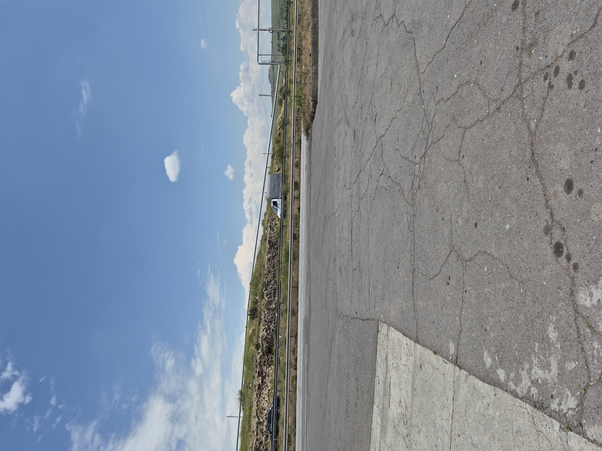 The image shows a clear blue sky with scattered white clouds. A multi-lane highway is visible in the middle ground, separated by a metal barrier. A black car is traveling on the highway on the left side of the image, and a white van with a covered bed is traveling on the highway on the right side. The highway is bordered by a grassy embankment with some rocky outcrops. In the foreground, there is a cracked asphalt road leading away from the viewer. The scene appears to be an outdoor setting in a semi-rural area, possibly on the outskirts of Ashtarak, Armenia, given the presence of a highway and a natural landscape. There are no people visible in the image.