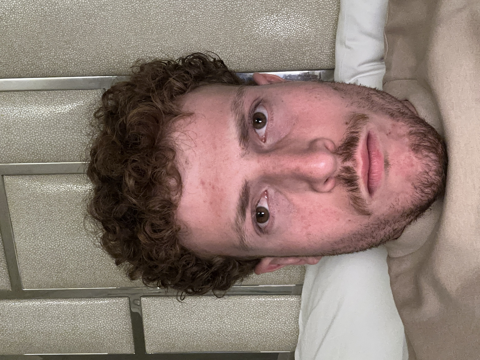 A close-up, slightly angled view shows a person's face. The individual has curly brown hair and a short beard. Their eyes are brown, and they appear to be looking directly at the camera. The person is wearing what seems to be a white t-shirt or garment, with the edge of a beige sweater or other piece of clothing visible beside it. The background behind the person features a wall with a textured pattern, comprised of rows of tiles, with a thin, horizontal silver bar accentuating each row. I cannot determine a specific location within Aventura, United States of America, based on the provided image.