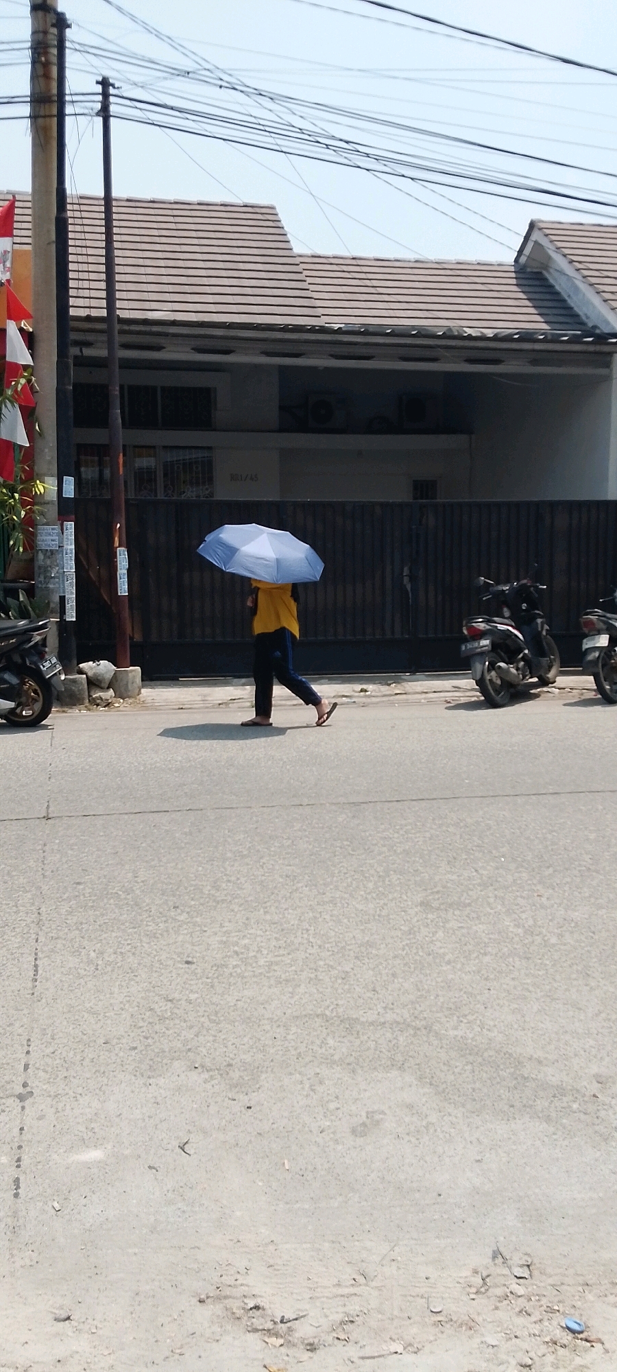 A person is walking on the side of a road in Satriamekar, Indonesia, holding a blue umbrella over their head for shade. The person is wearing a yellow shirt and dark pants. They are walking from left to right in the frame. Several motorcycles are parked on the side of the road. A house with a tiled roof is visible in the background. Overhead, a tangle of power lines crisscrosses the sky. The ground is paved and appears to be a road surface.